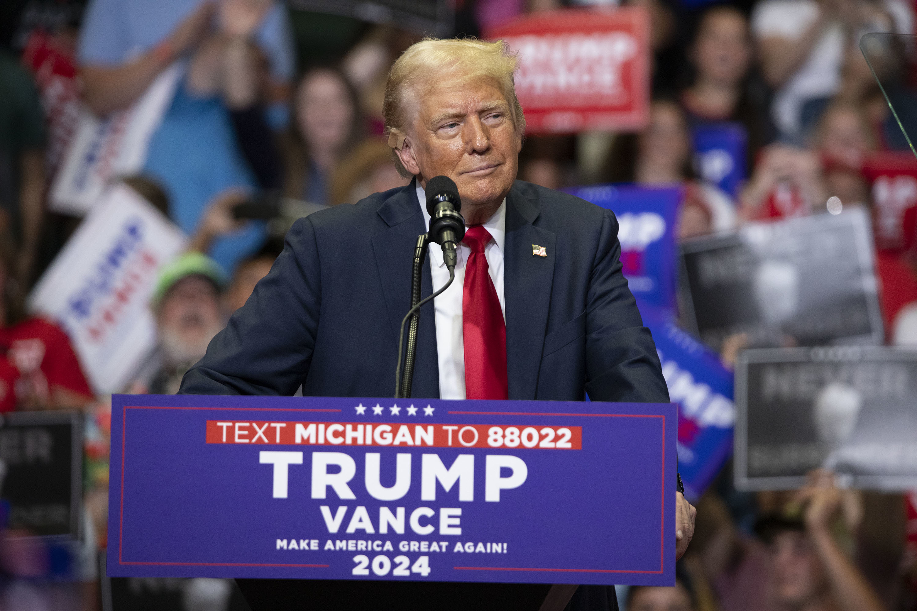 Donald Trump speaking at a rally, standing behind a podium with a sign that reads "Trump Vance 2024." Audience members hold signs and banners in the background