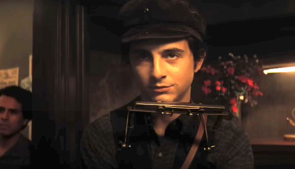 A young man in a dark cap and striped shirt stands holding a harmonica. There is a person and flowers in the dimly lit background