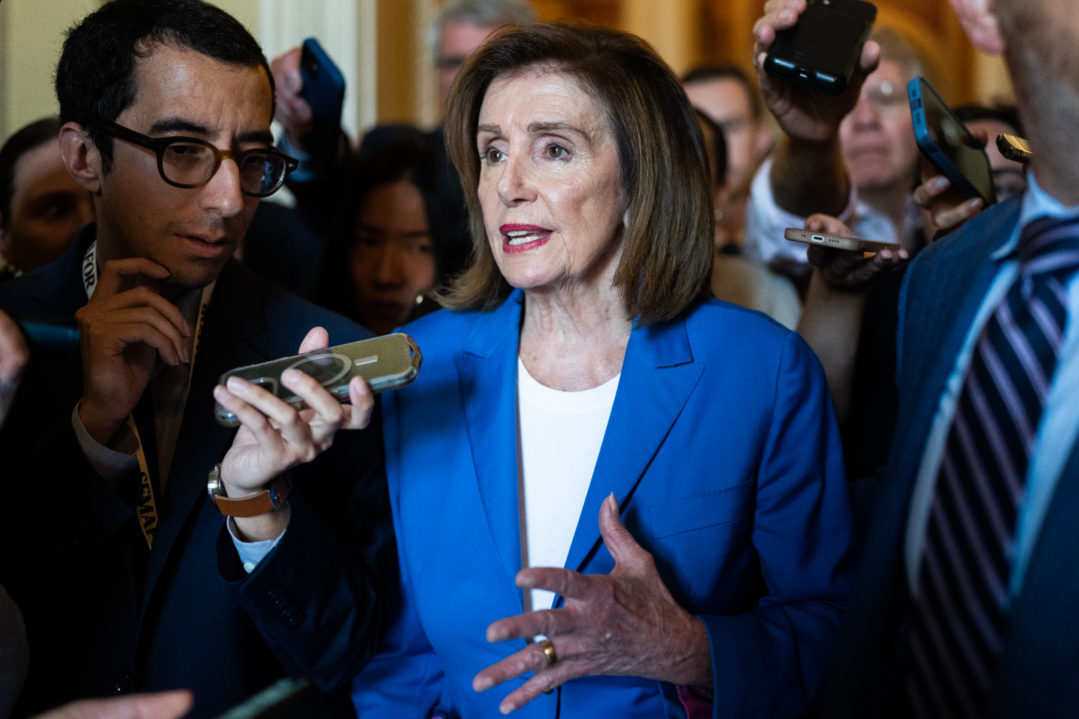 Nancy Pelosi speaks to reporters, surrounded by people holding microphones and recording devices. She is wearing a blue blazer over a white top