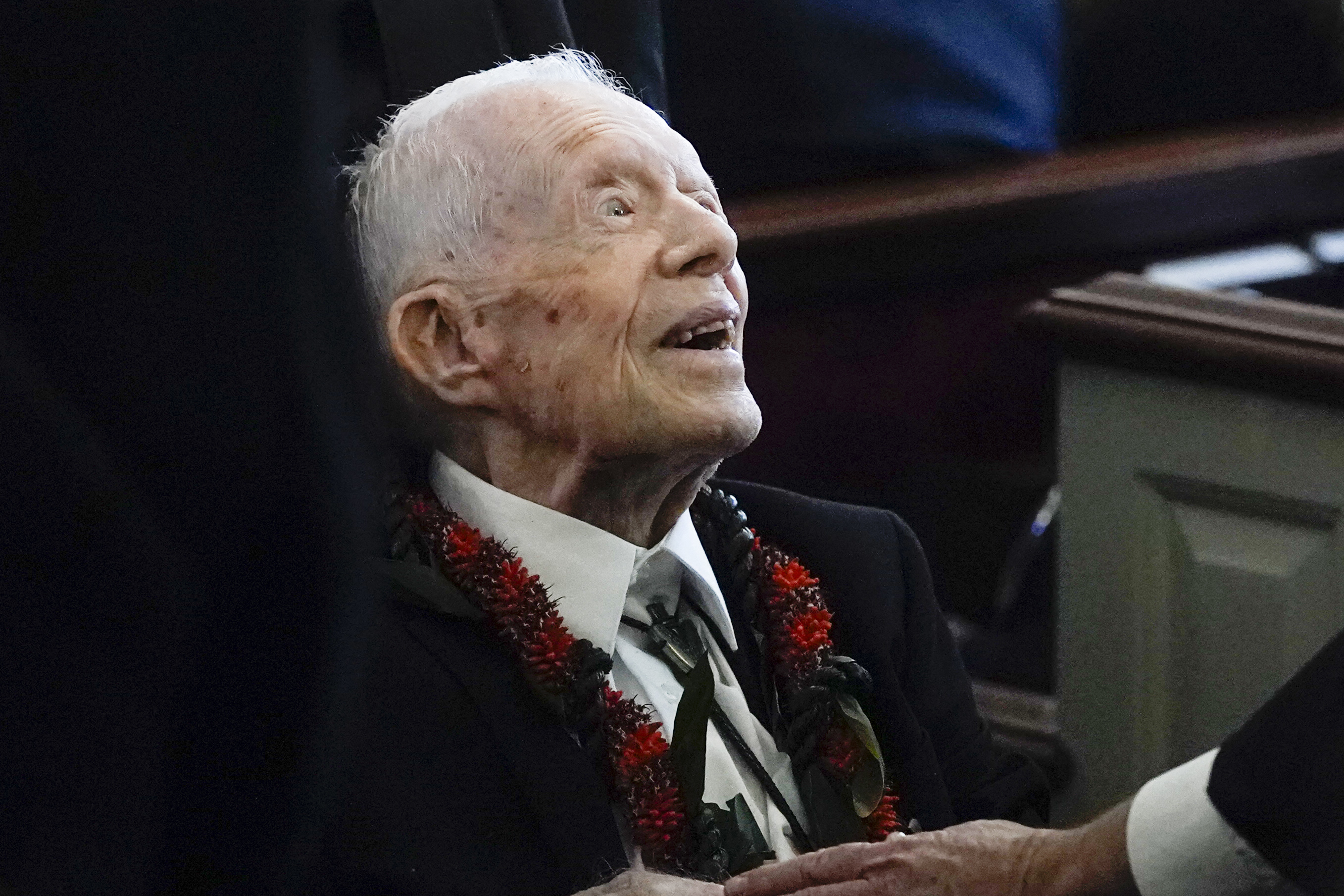 Jimmy Carter, wearing a suit and floral lei, looks upward with a pleased expression while seated indoors