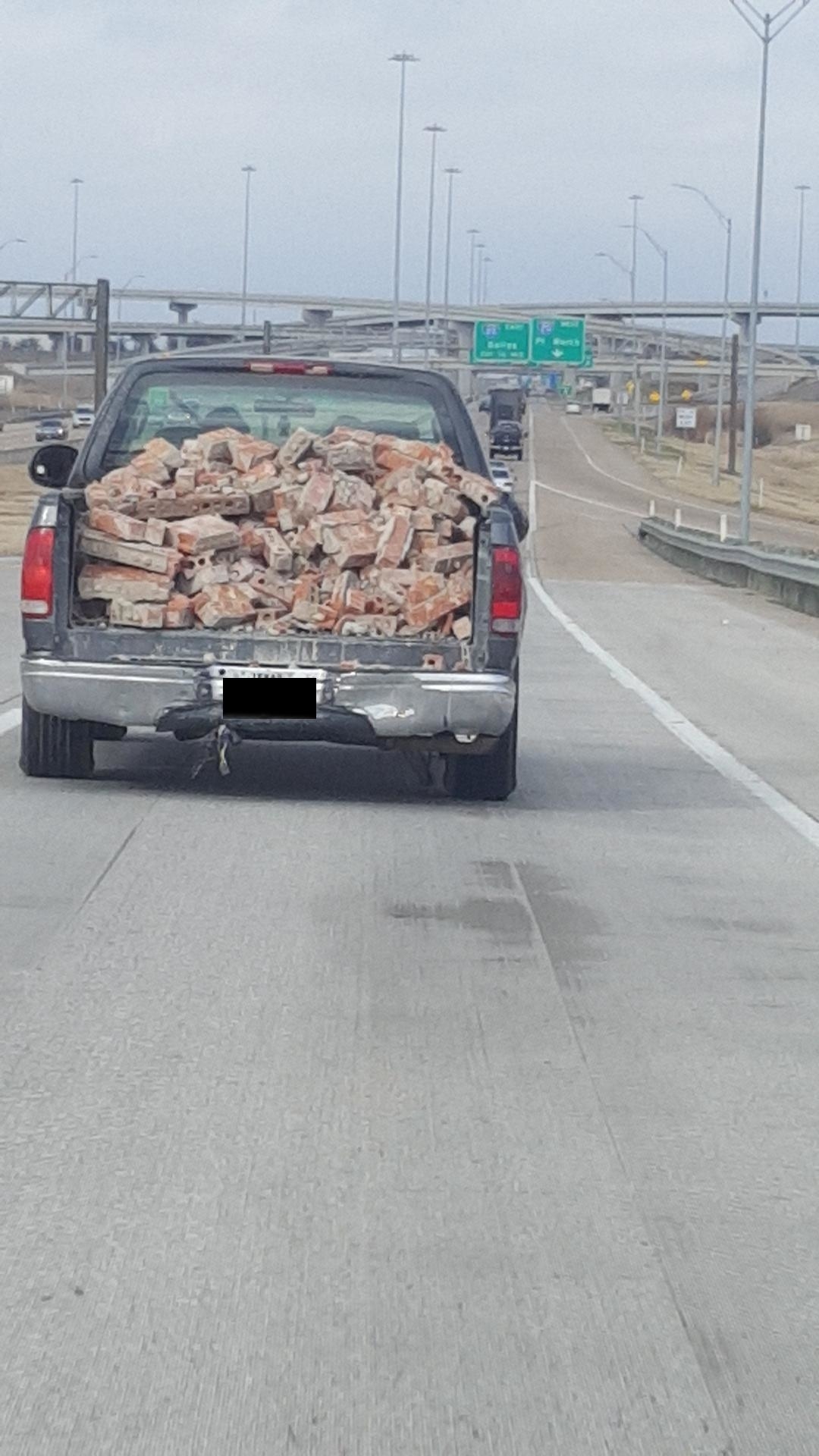A pickup truck loaded with bricks drives on a highway. The truck's tailgate is down, and the rear bumper is dented