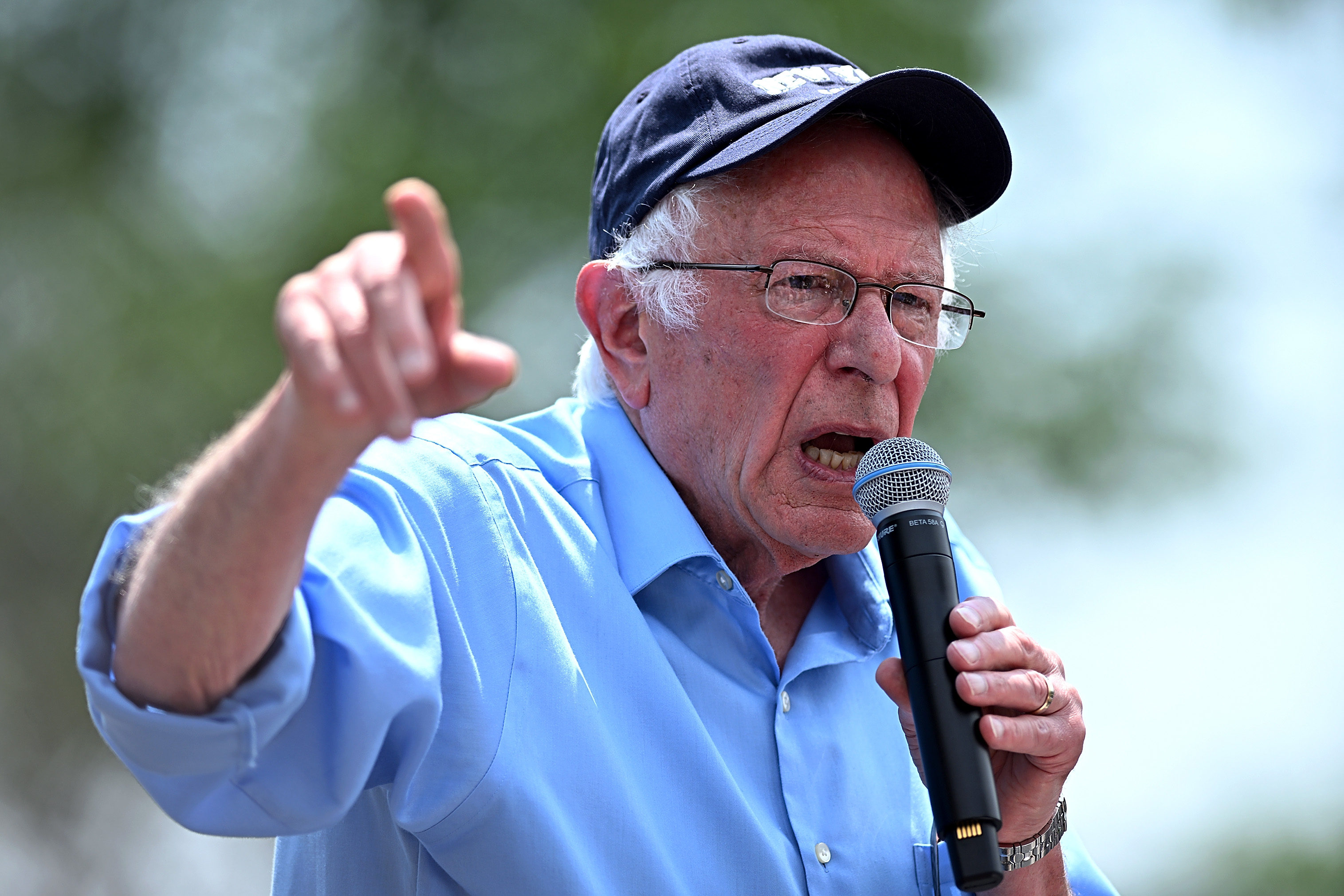 Bernie Sanders speaks passionately into a microphone, wearing a casual blue button-up shirt and a dark cap. He gestures with his other hand
