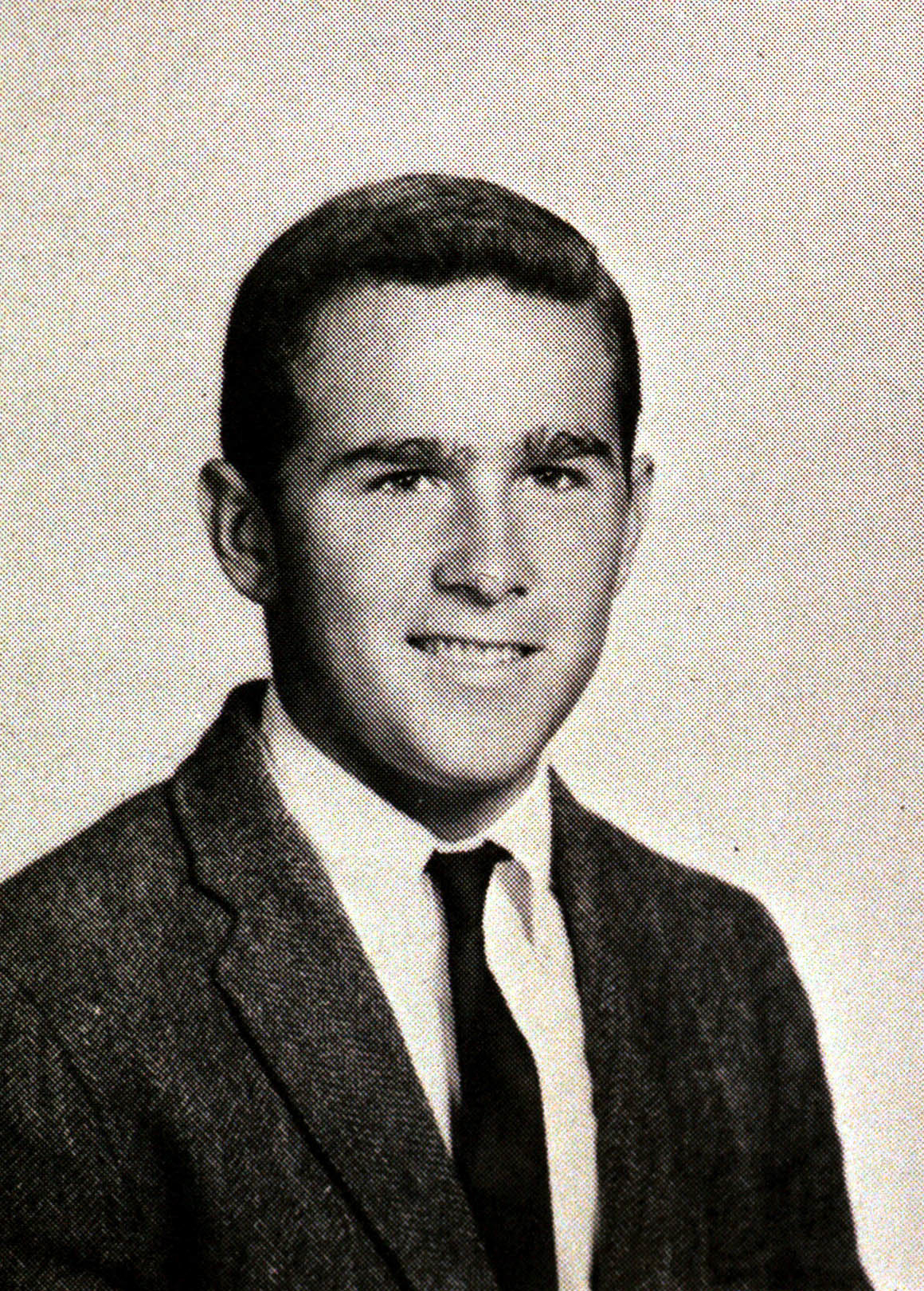 A young man wearing a suit jacket, dress shirt, and tie, smiling at the camera in a formal portrait