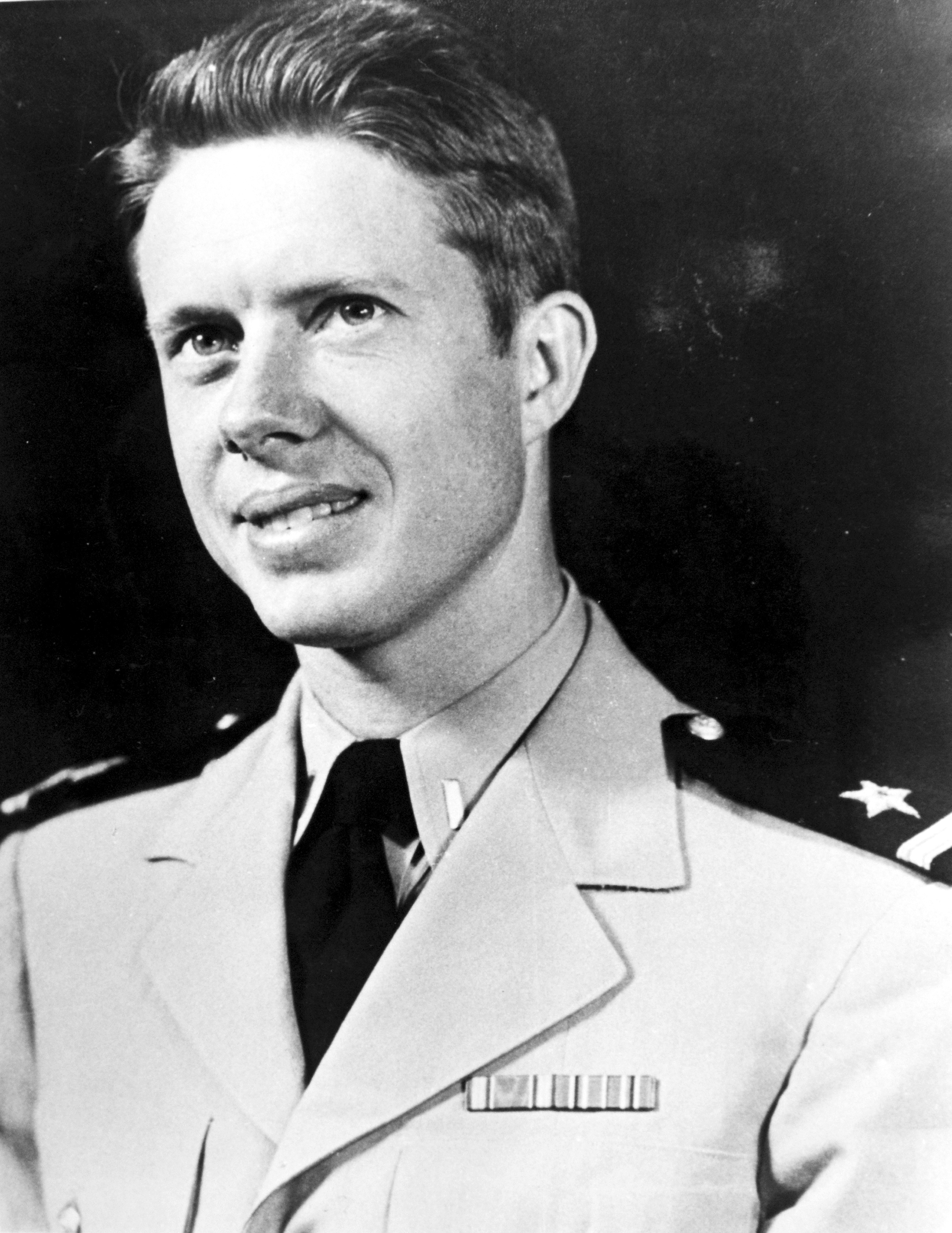 A young man in a military uniform with badges and insignia smiles slightly. His hair is neatly combed. The background is plain and dark