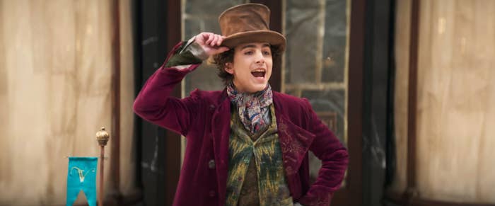 Timothée Chalamet, wearing a top hat and ornate Victorian-style clothing, poses with a hand on his hat in front of a classic building