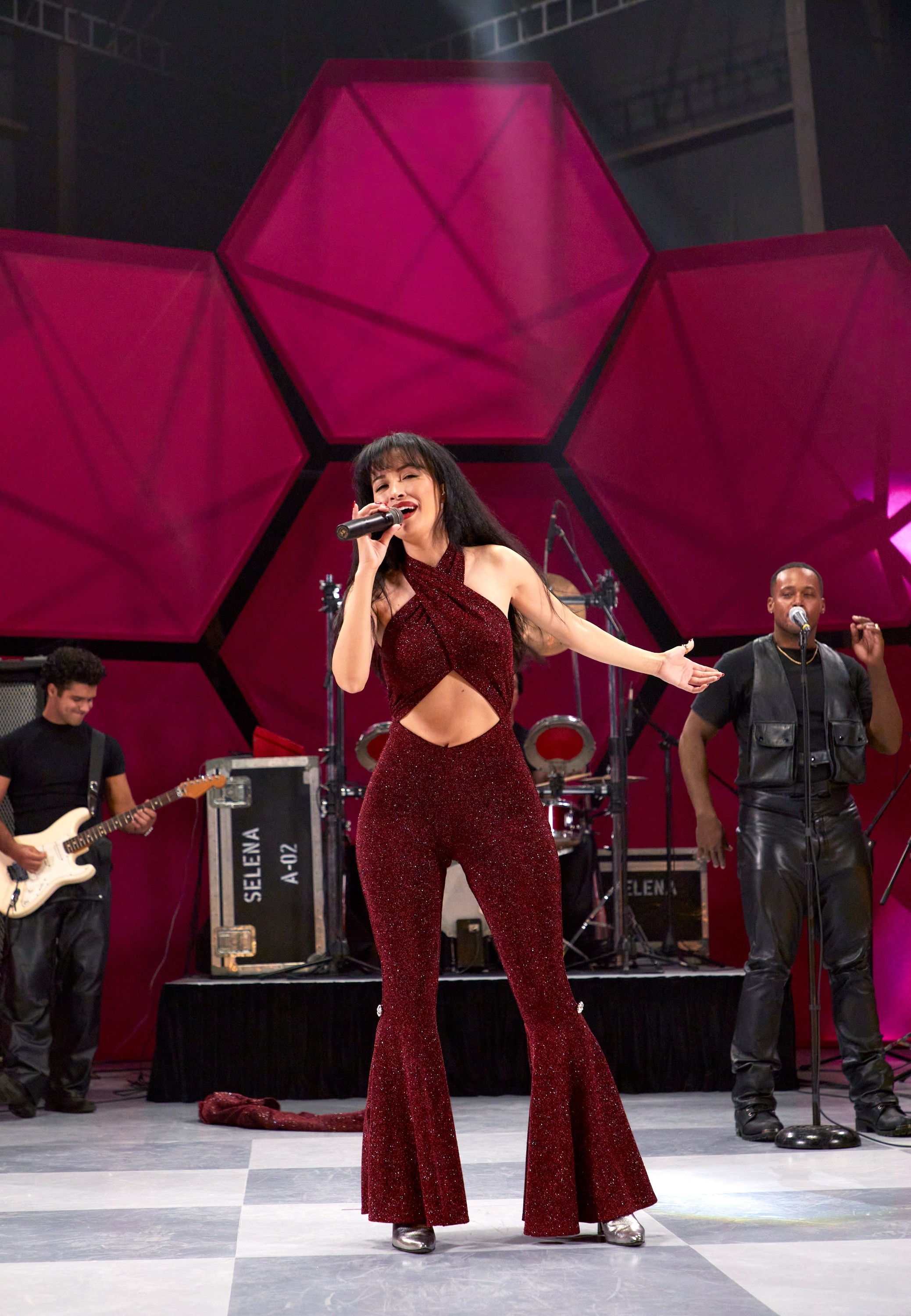 Selena Quintanilla dancing and singing on stage in a sparkling jumpsuit while musicians play instruments behind her