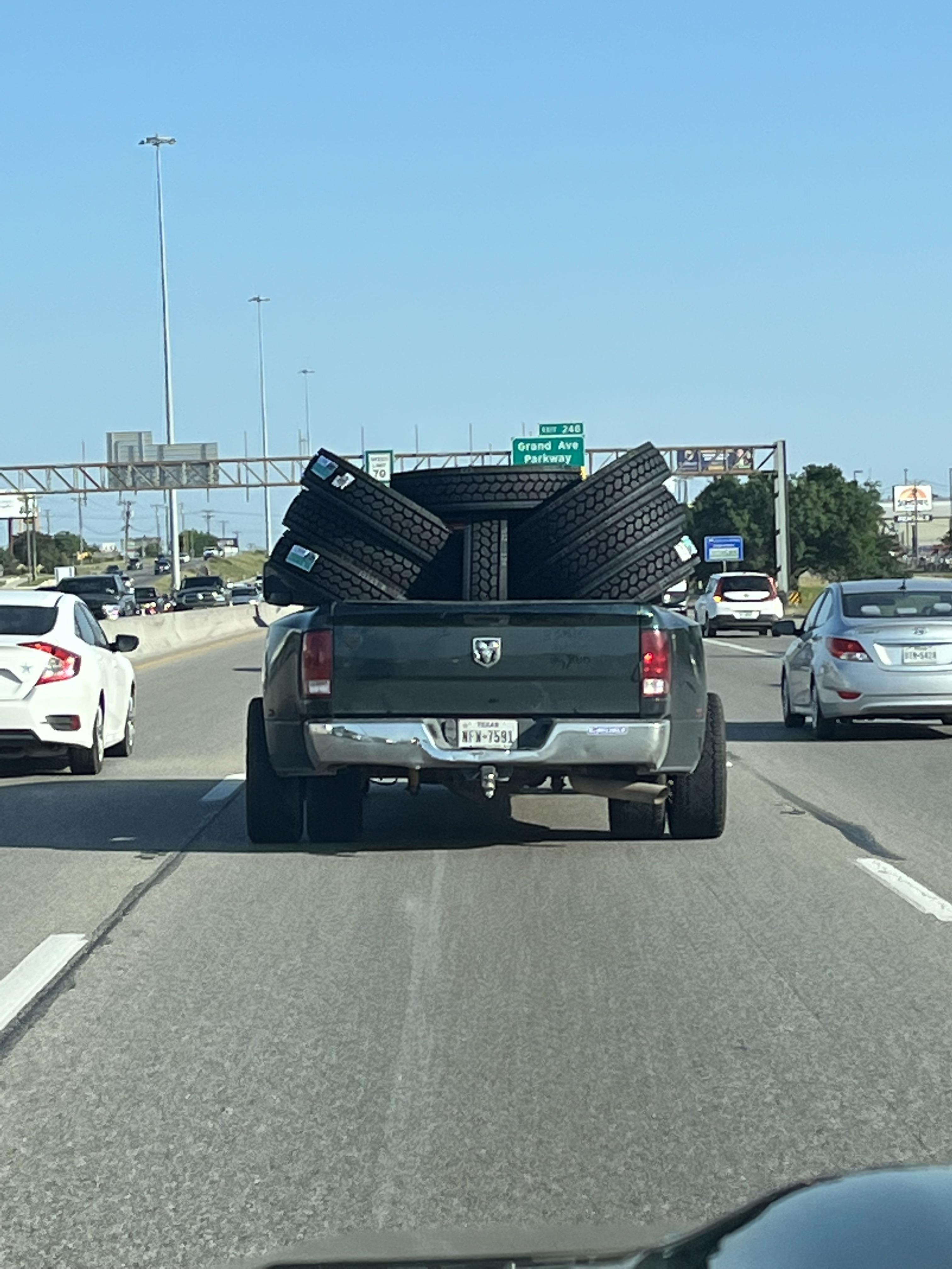 A pickup truck loaded with multiple tires in its bed is driving on a highway, flanked by other cars. Road signs and trees are visible ahead