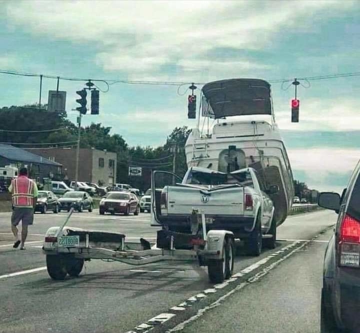 A pickup truck towing a large boat on a trailer is stuck in the middle of a busy intersection. A pedestrian walks nearby under traffic lights