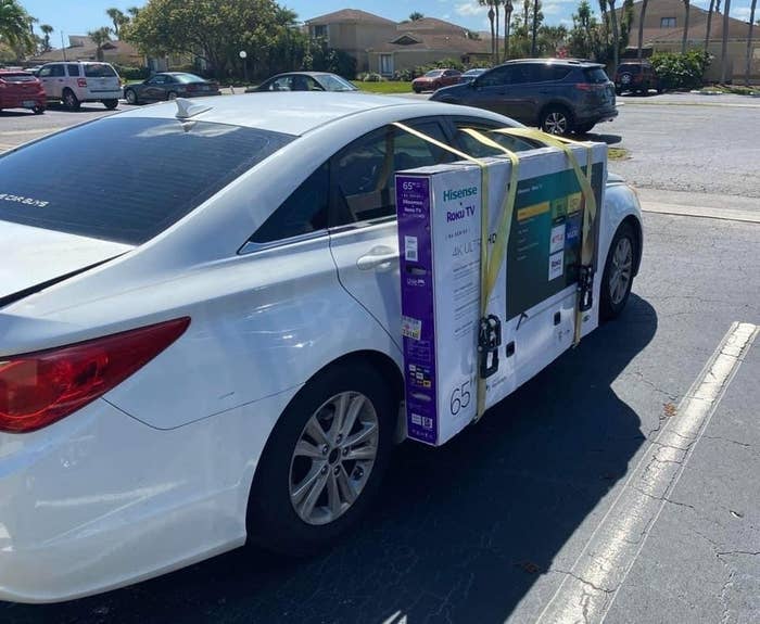 A large Hisense Roku TV box is strapped to the side of a white car with yellow straps in a parking lot