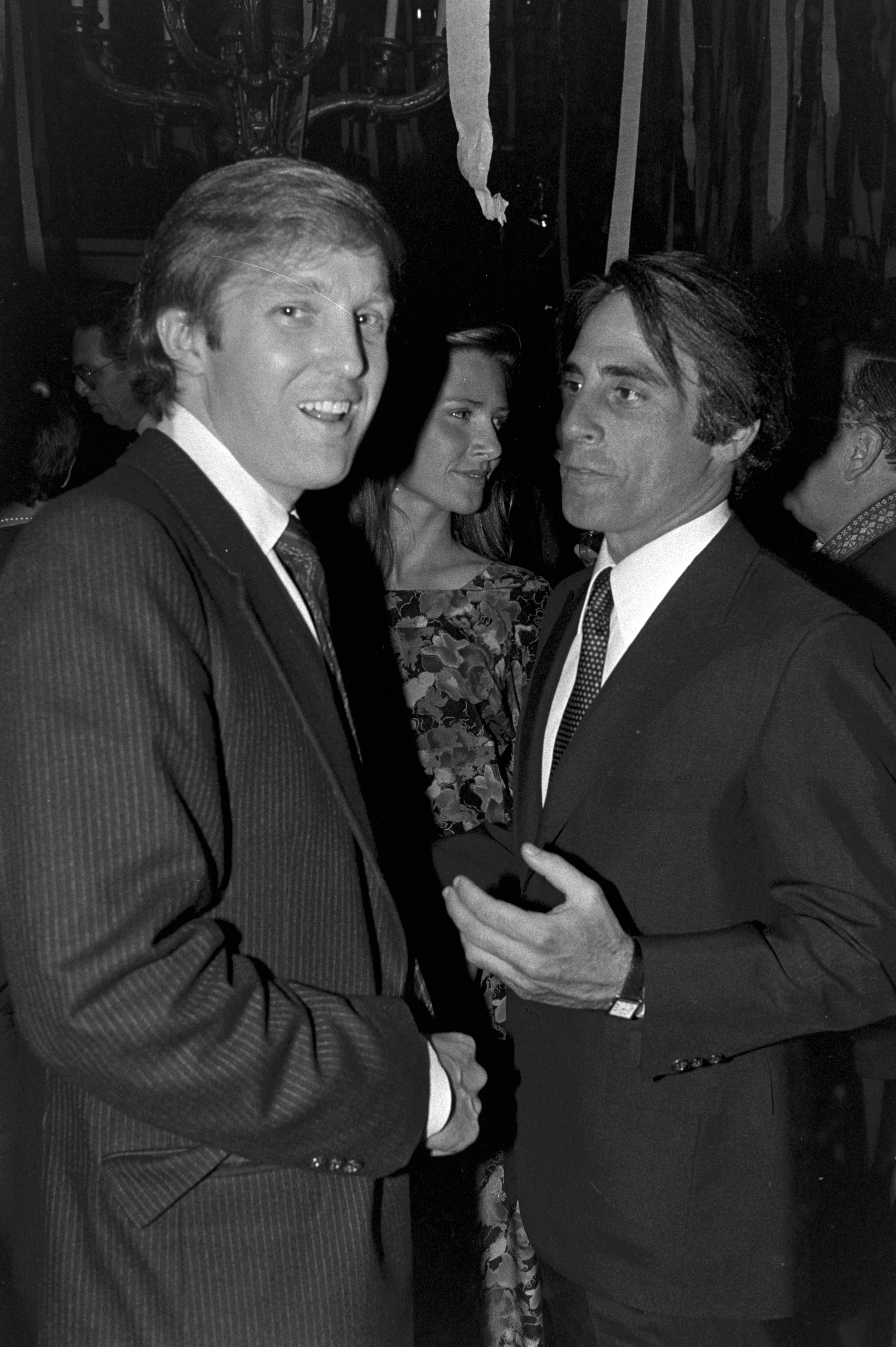 Donald Trump and another person in suits, talking at an event. A woman stands in the background