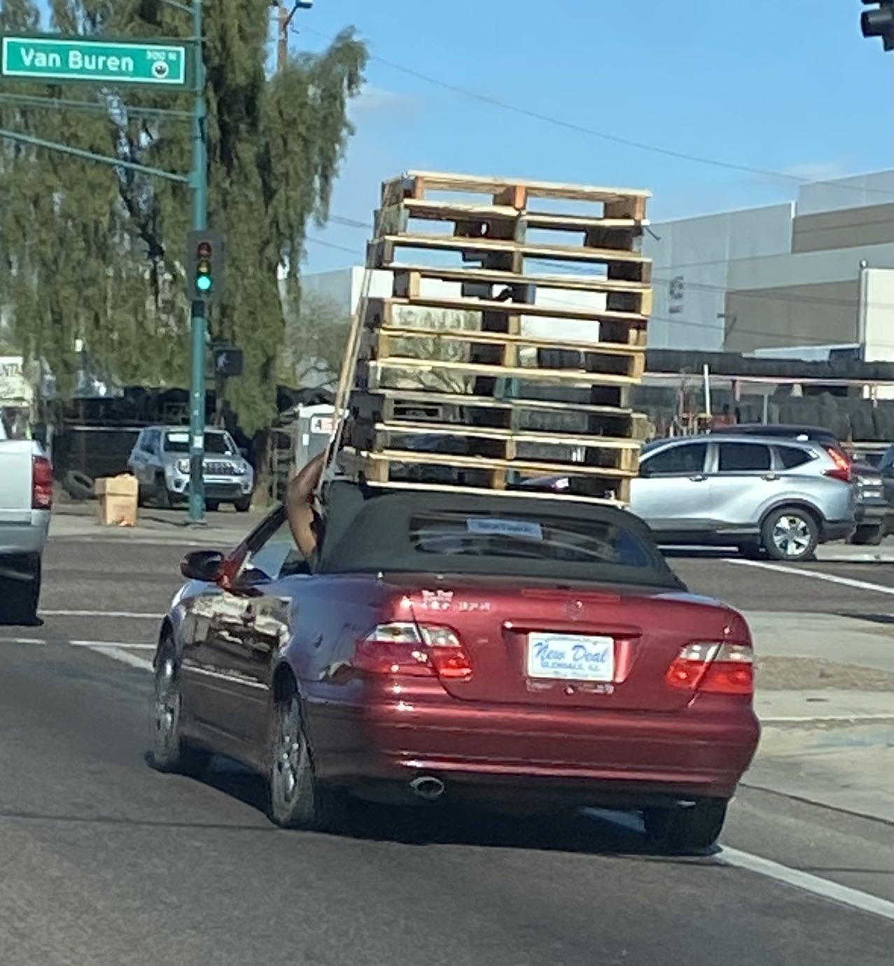A red convertible car with a large stack of wooden pallets held by the driver is seen on a busy street