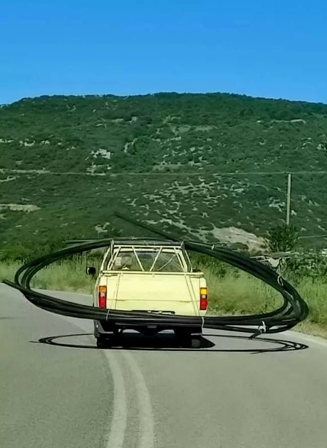 A pickup truck is driving on a road with large, circular pipes awkwardly loaded and extending out from both sides of the vehicle