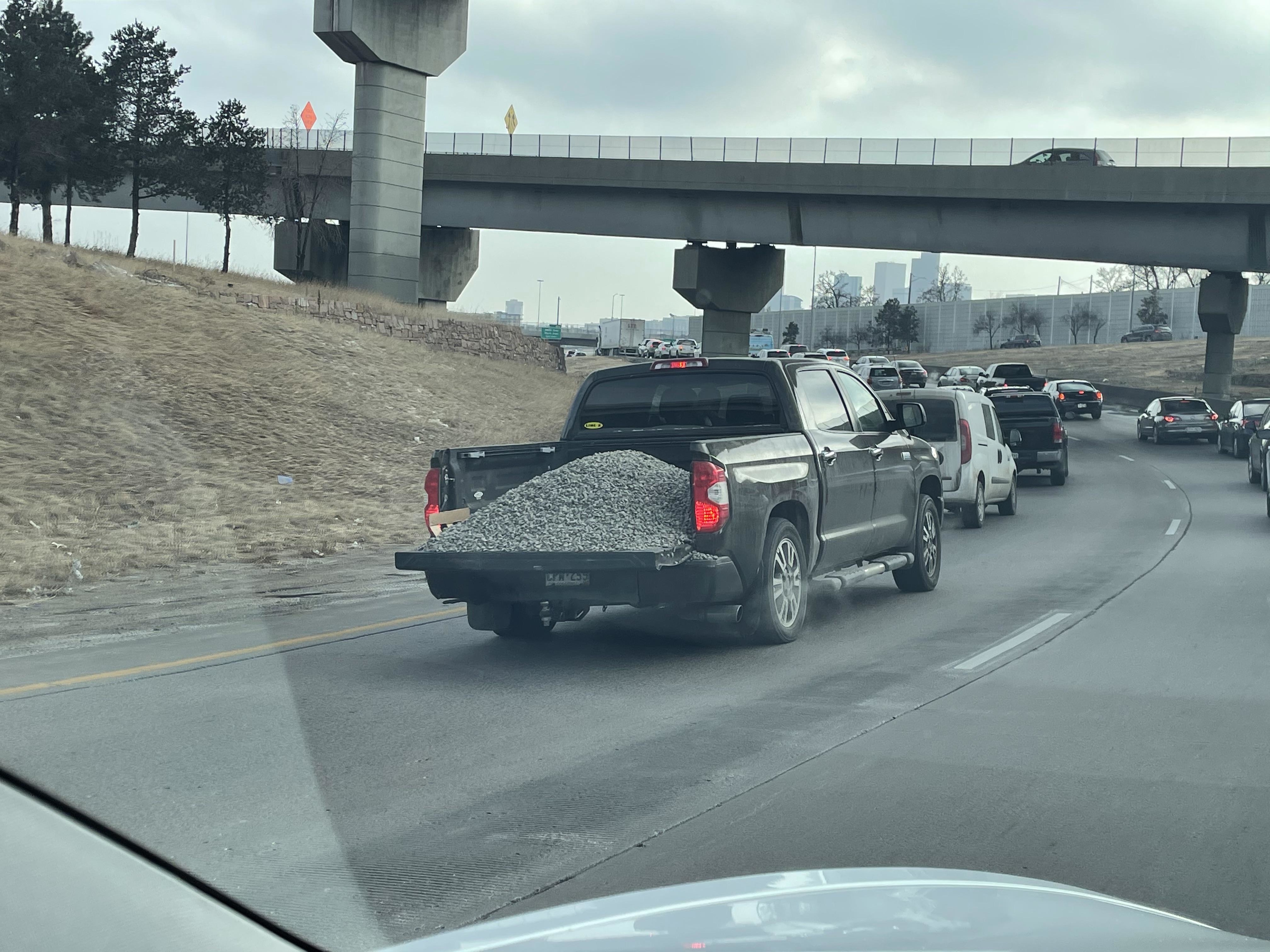Pickup truck filled with gravel driving in heavy traffic on a highway beneath an overpass. Other cars are visible in the lanes