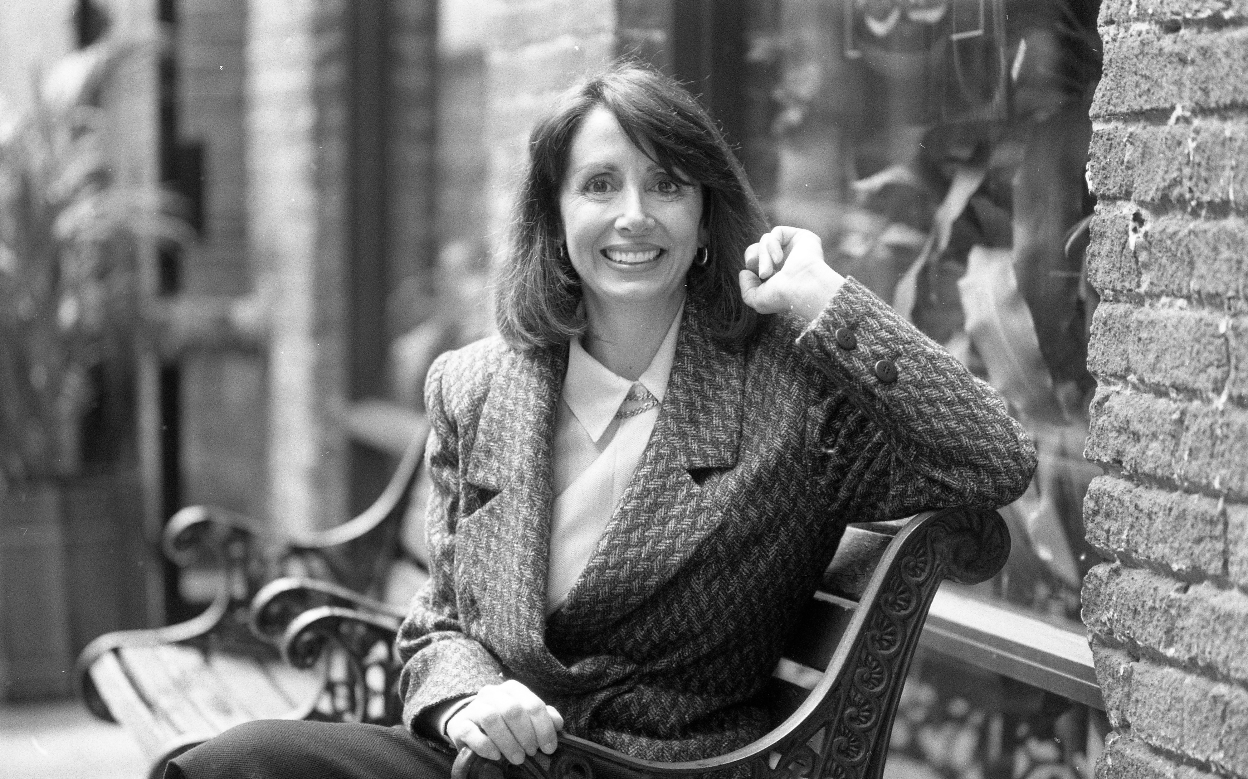 A woman in a houndstooth blazer smiles while sitting on a bench outside a building with brick walls and large windows