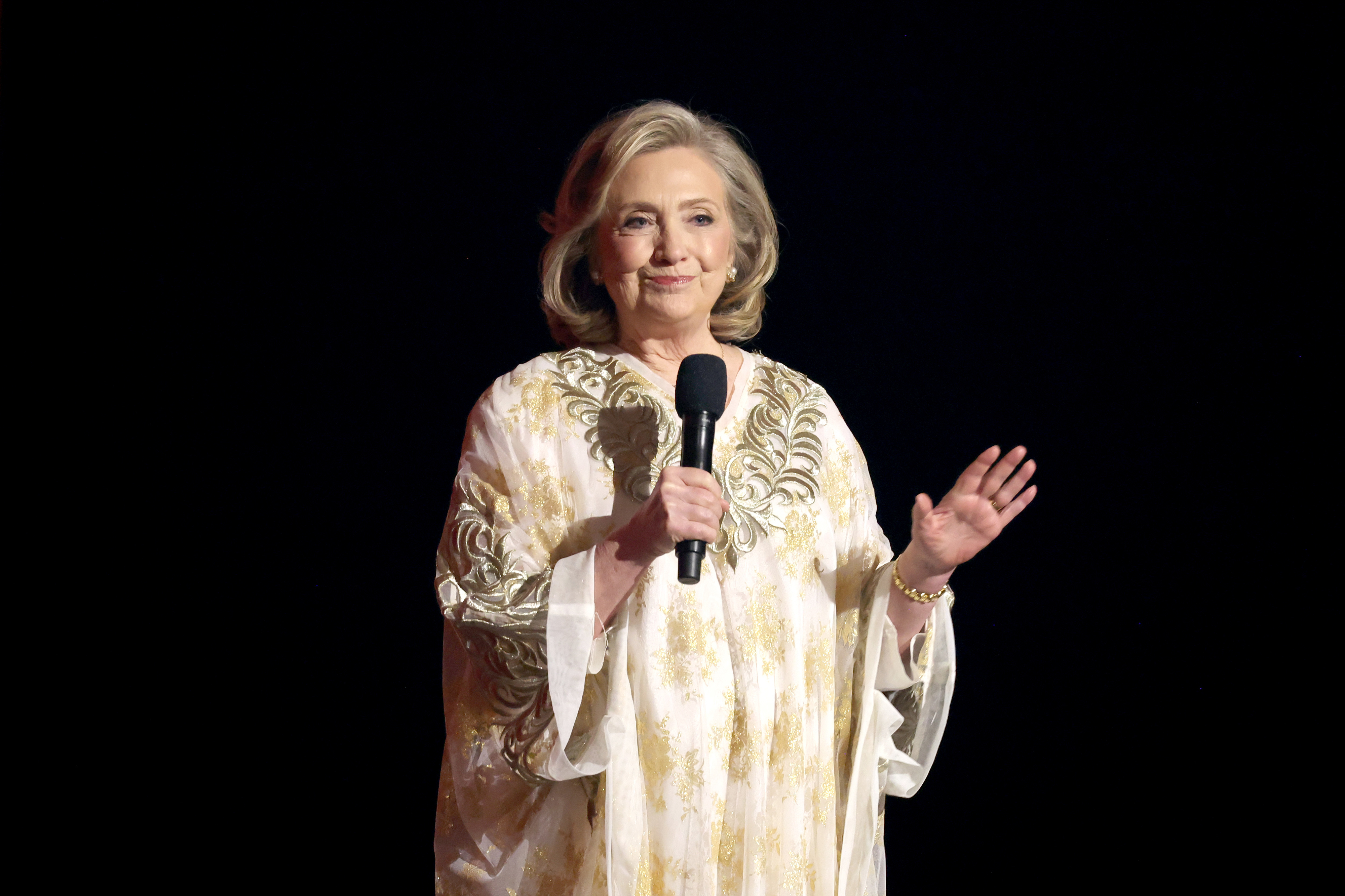 Hillary Clinton is standing on stage in a flowing patterned dress, holding a microphone and gesturing with her other hand