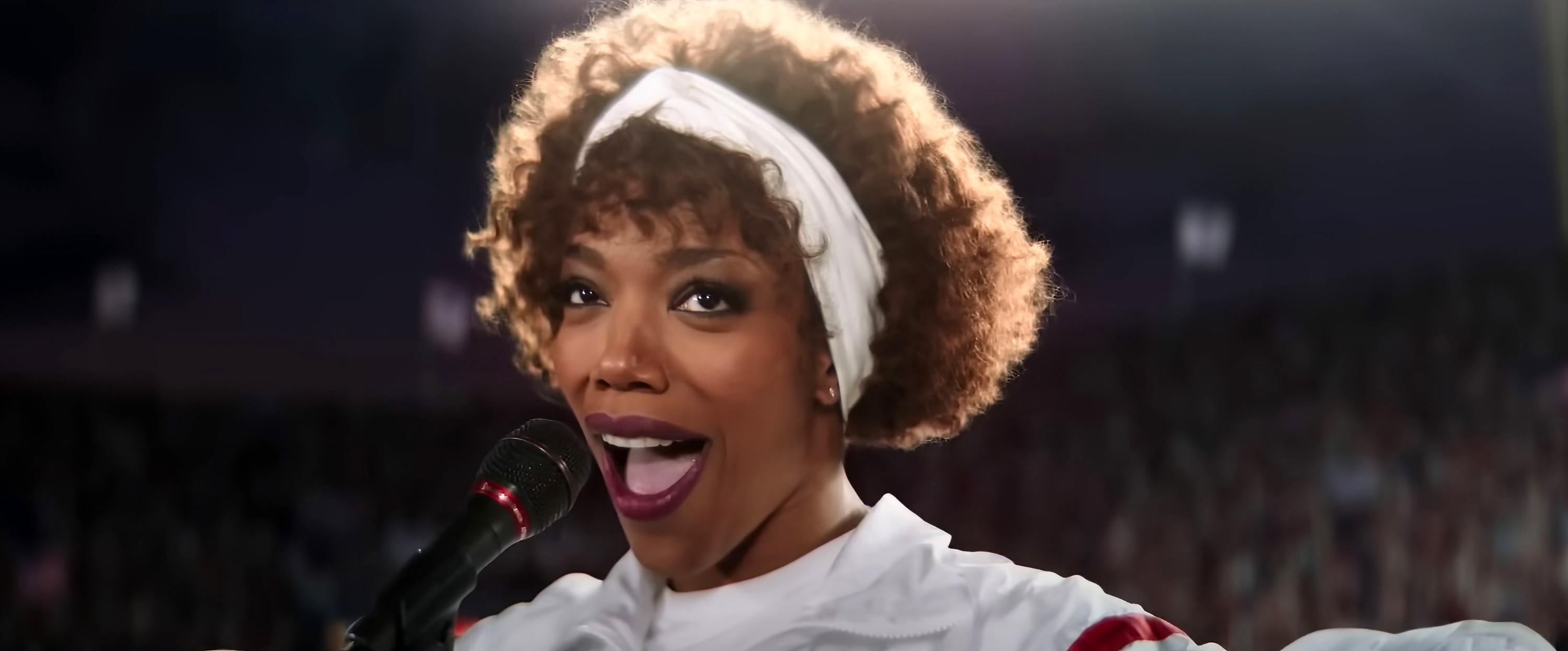 Anika Noni Rose singing into a microphone, wearing a white headband and a white top with a red stripe on the sleeve