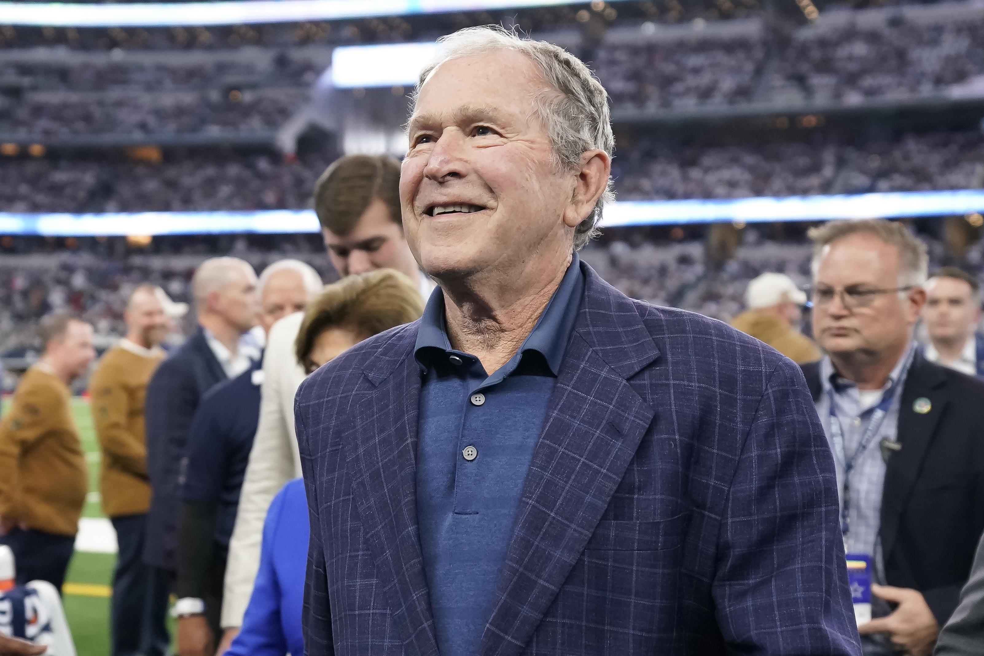 George W. Bush in a blue shirt and checked blazer, smiling at an event with a crowd in the background