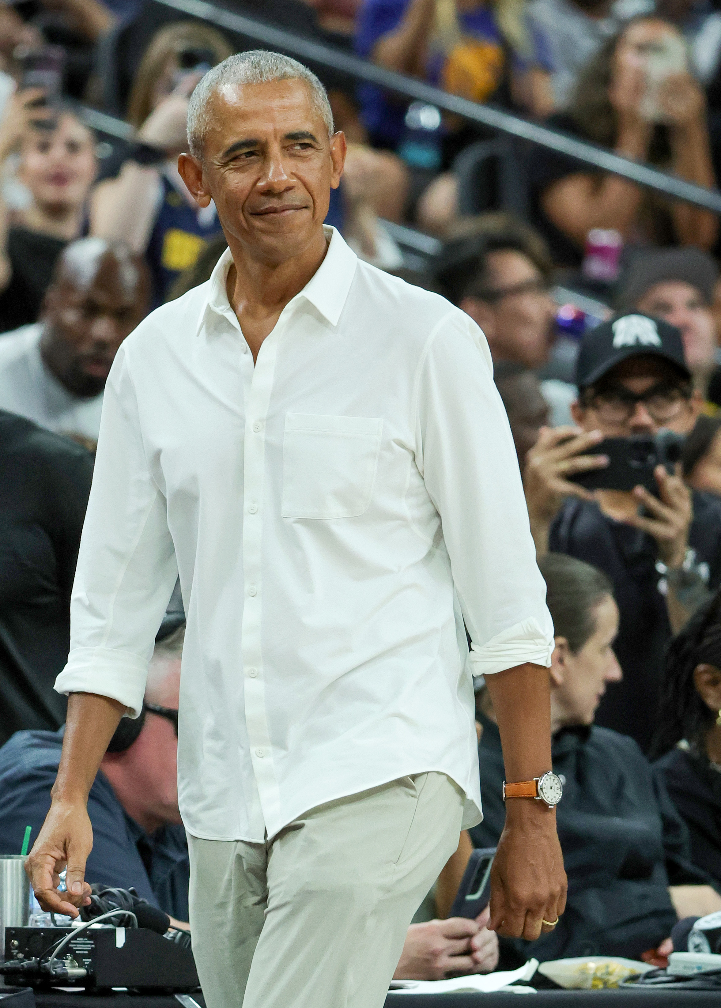 Barack Obama is seen walking in a crowd, wearing a casual white shirt and beige pants. People in the background are taking photos and watching him