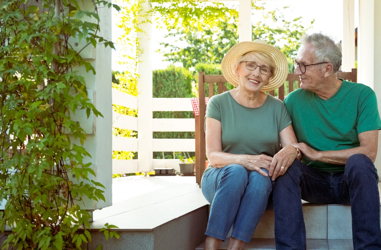 An elderly couple sits on porch steps holding hands and smiling, dressed casually