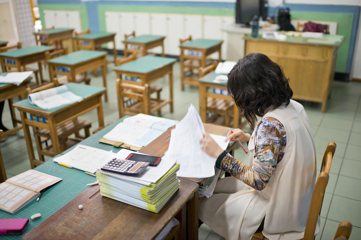 A woman grades papers at a desk in an empty classroom