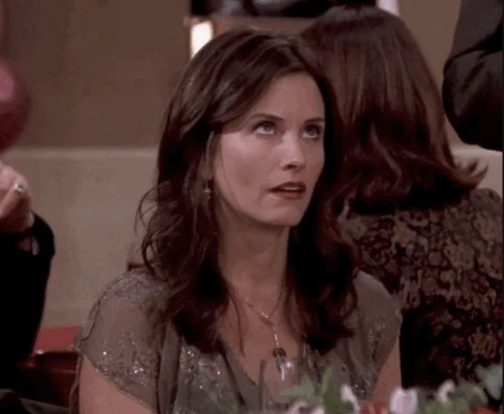 Courteney Cox, with an exasperated expression, is seated at a dining table in a formal setting