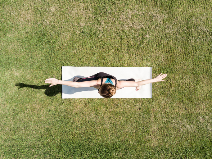 Person doing yoga in a seated pose on a white mat on a grassy field, with arms extended sideways