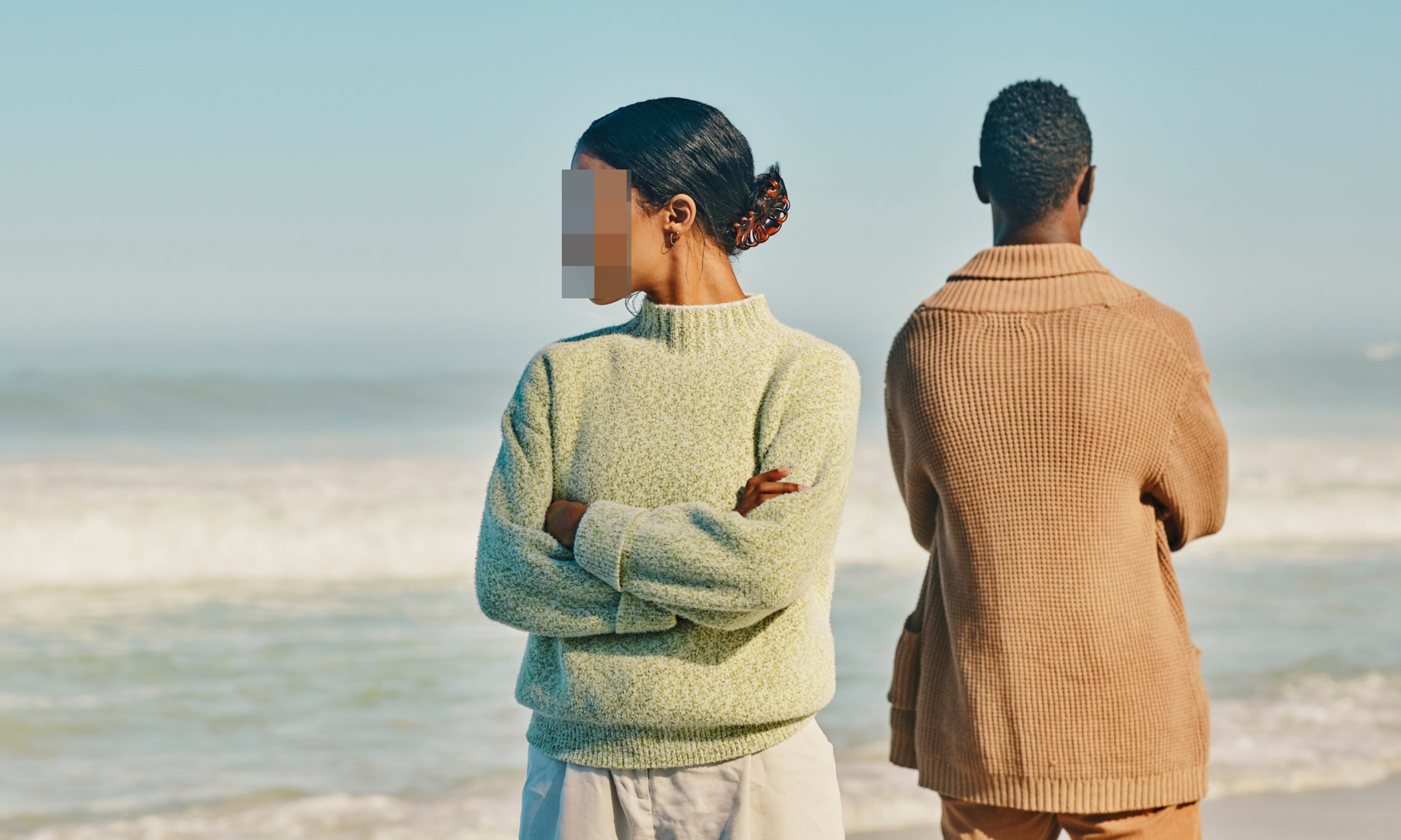 A woman and a man stand back-to-back on a beach, both with arms crossed, looking away from each other