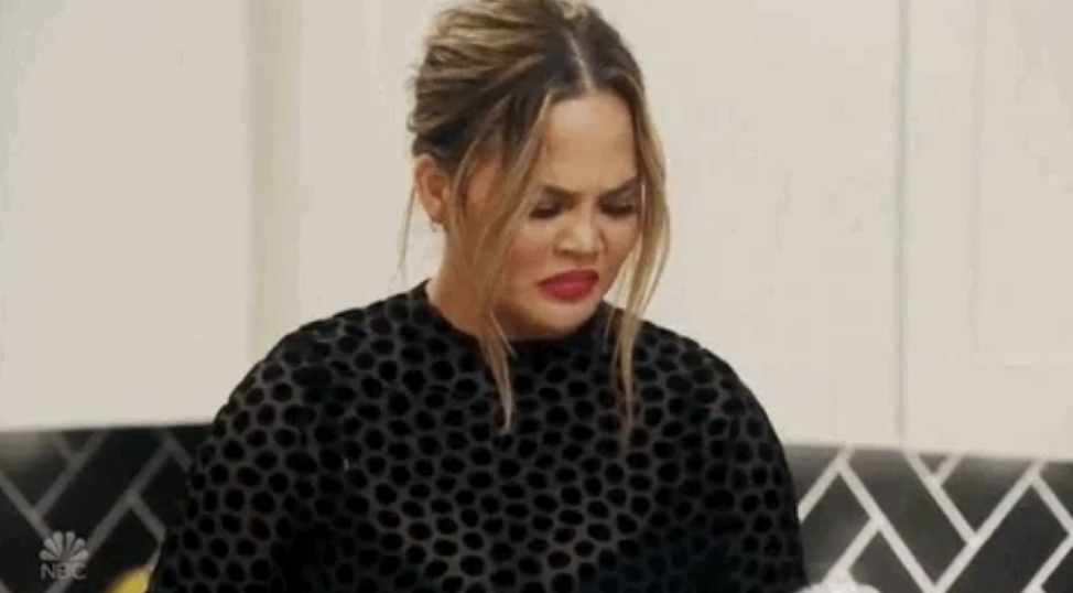 Chrissy Teigen making a disgusted or confused facial expression while looking at something off-camera. She is wearing a black, polka-dot top
