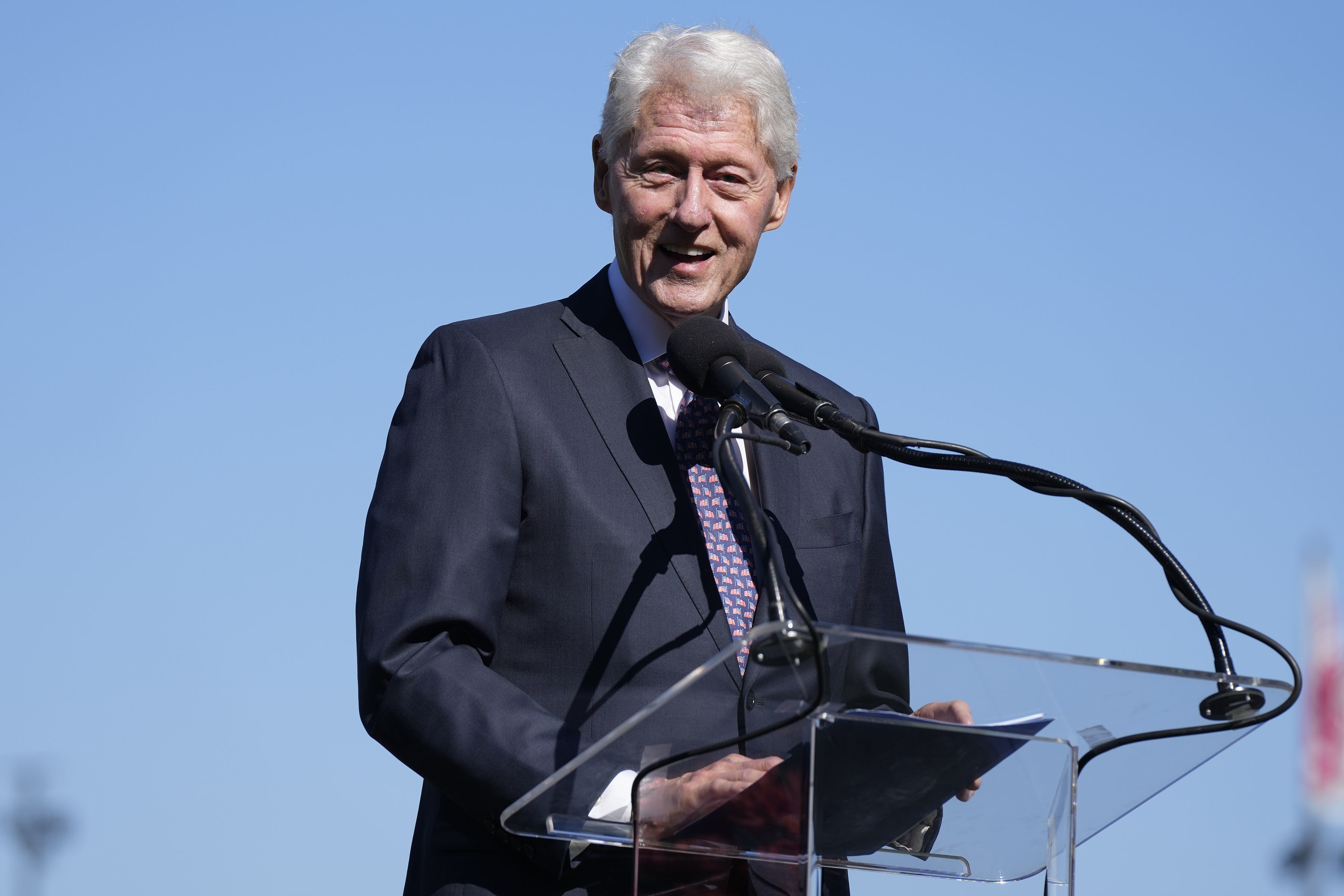 Bill Clinton speaks at a podium outdoors, holding a folder, with microphones in front of him