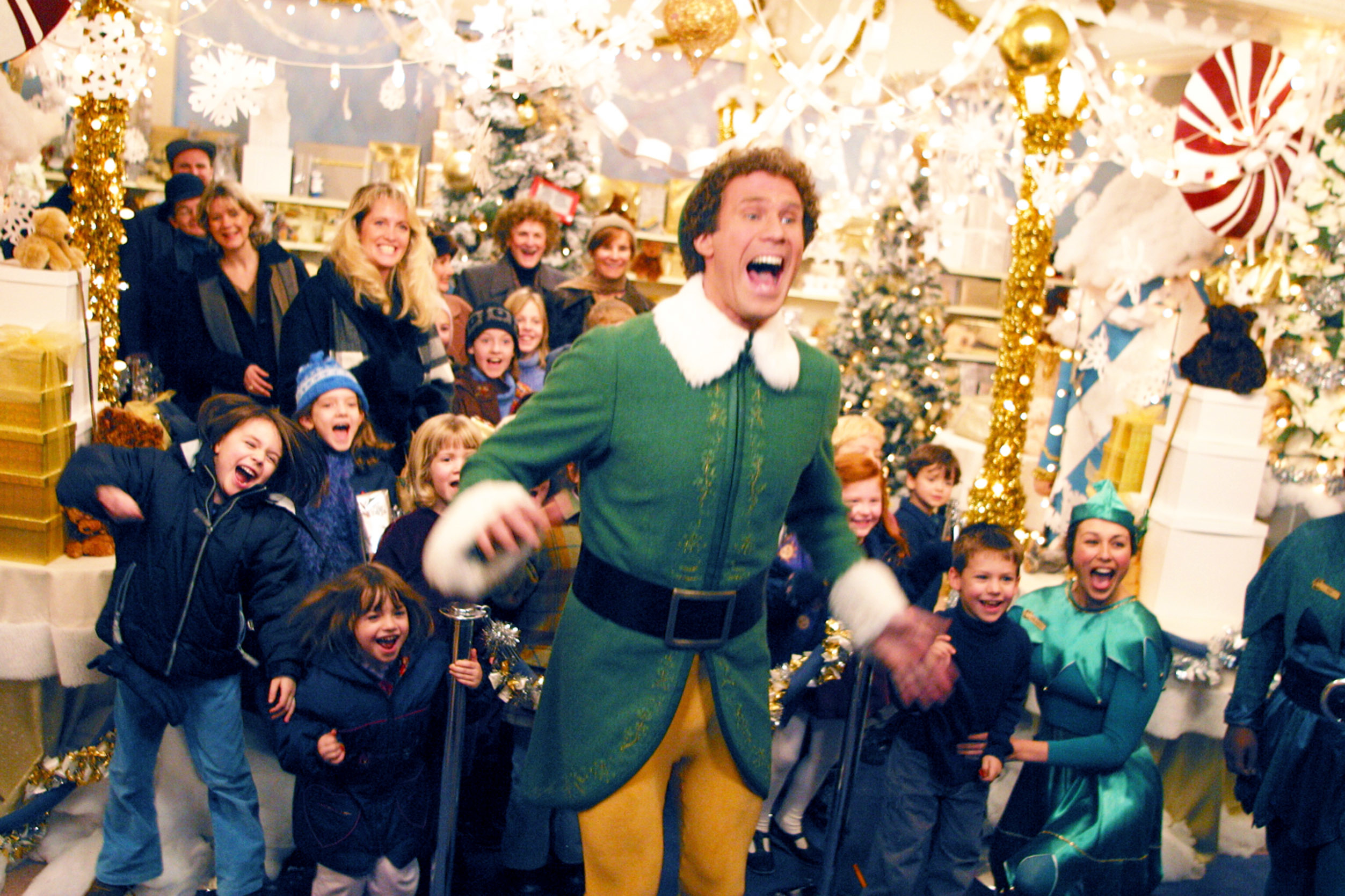 Will Ferrell, dressed as Buddy the Elf, joyfully leads a group of children and adults through a lively, festive Christmas display