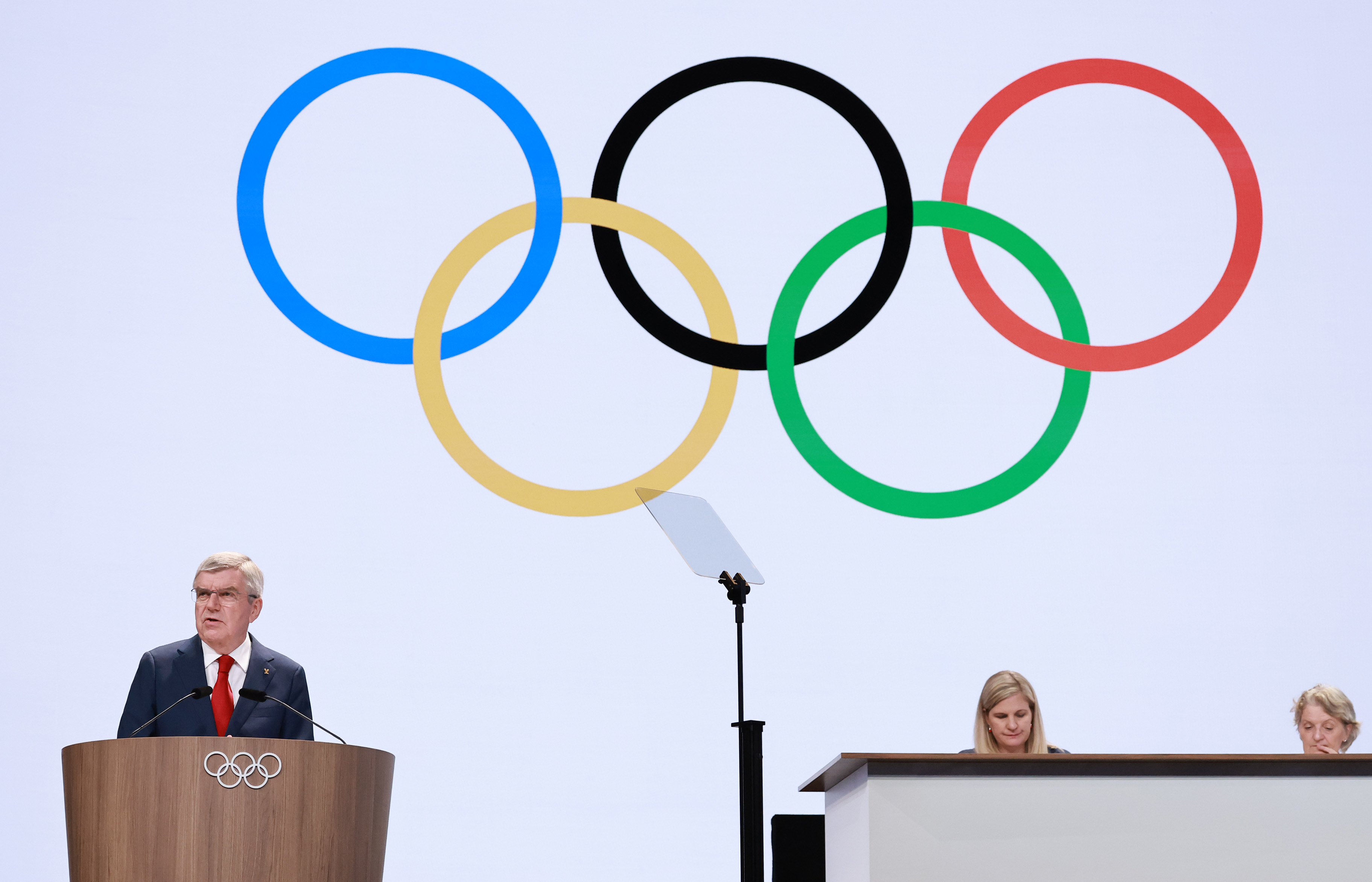 Thomas Bach speaks at a podium with the Olympic rings behind him, while two people sit at a table in the background