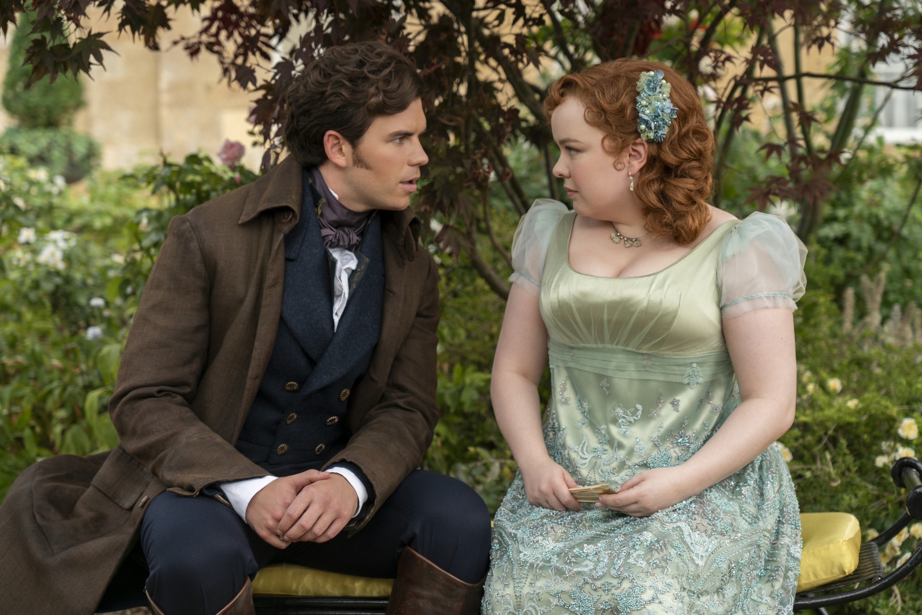 Luke Newton and Nicola Coughlan in period costumes, sitting on a bench and engaging in a conversation in a garden setting