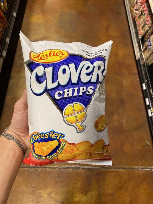A hand holding a bag of Leslie's Clover Chips Cheezy flavored corn snack in a grocery store aisle