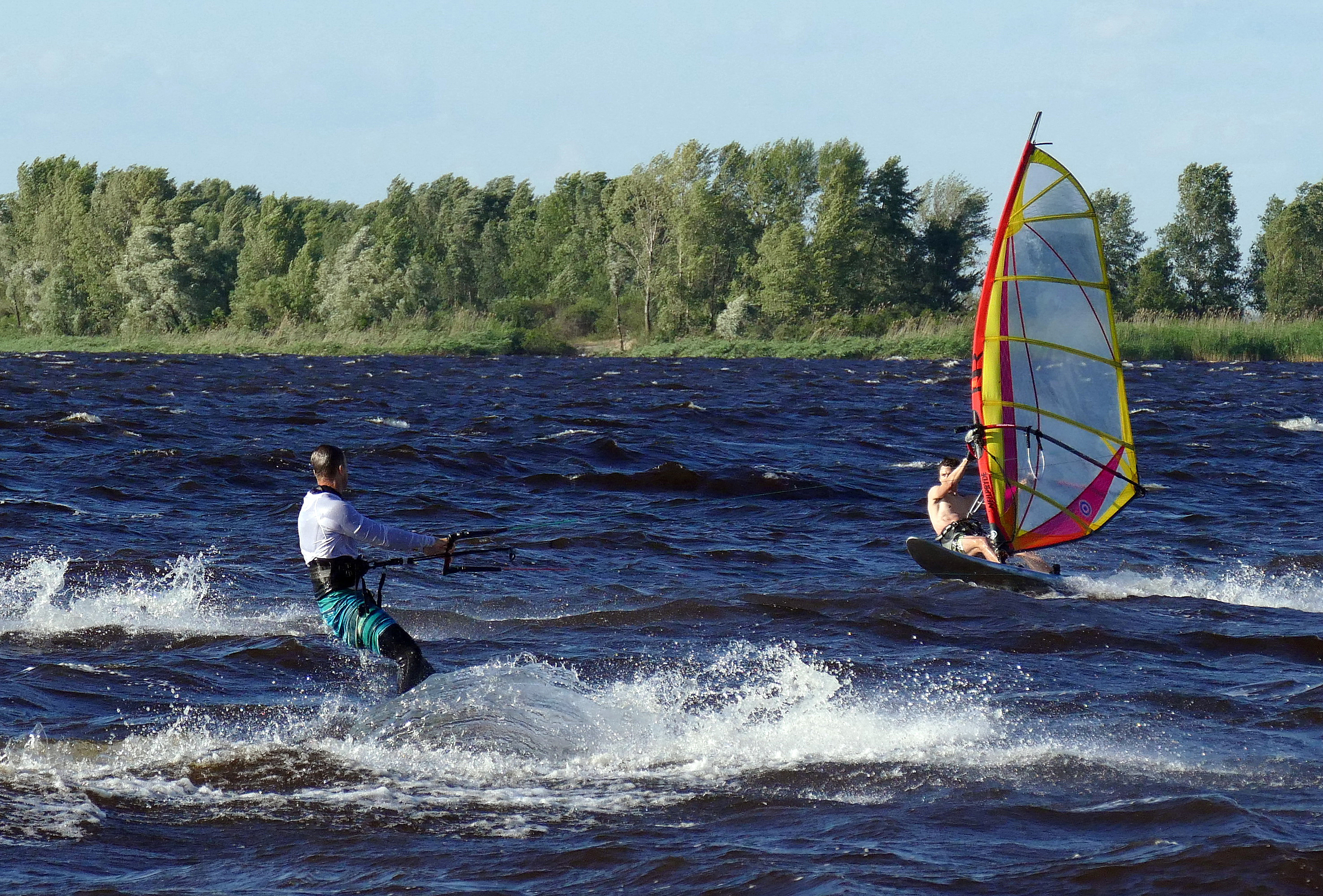 Two people engaging in water sports on a windy lake. One is kite surfing, and the other is windsurfing. Lush green trees line the background