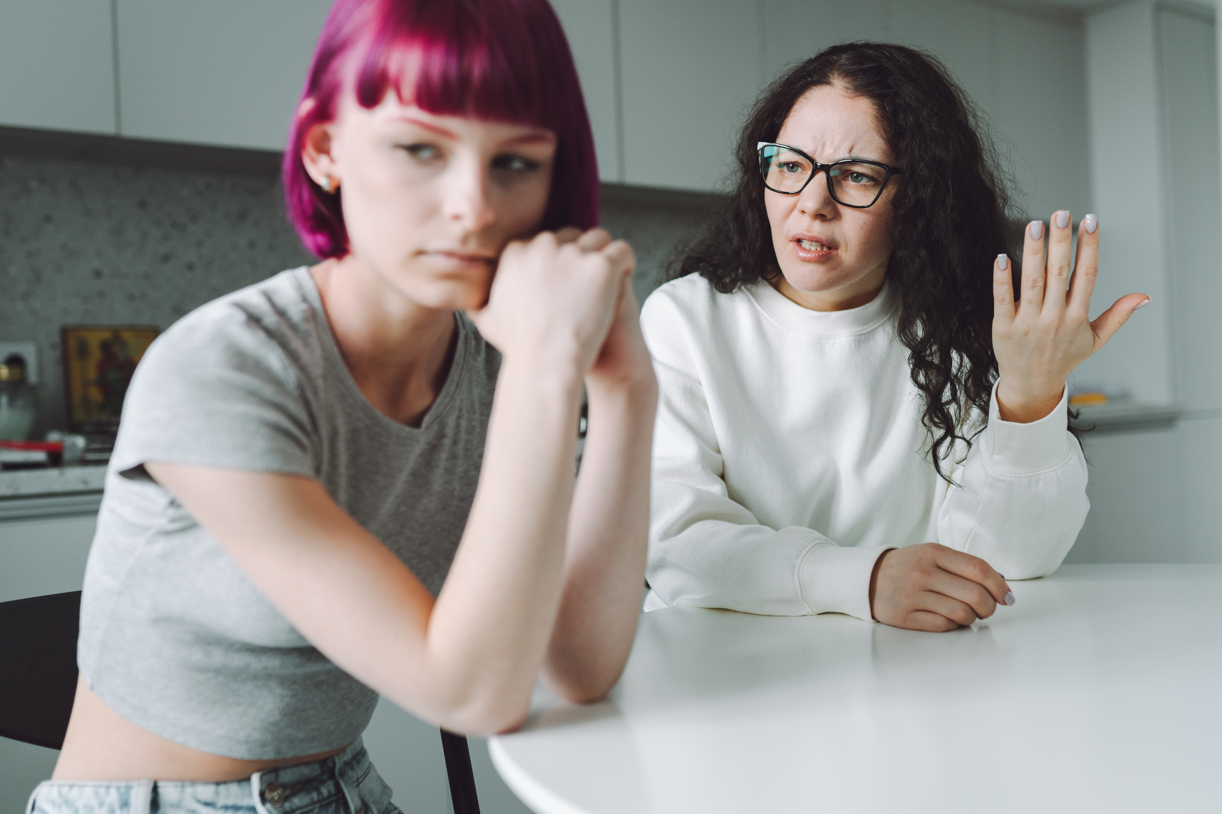 A mother and teen talking at a kitchen table. The teen with pink hair looks upset; the mother, with curly hair and glasses, appears concerned