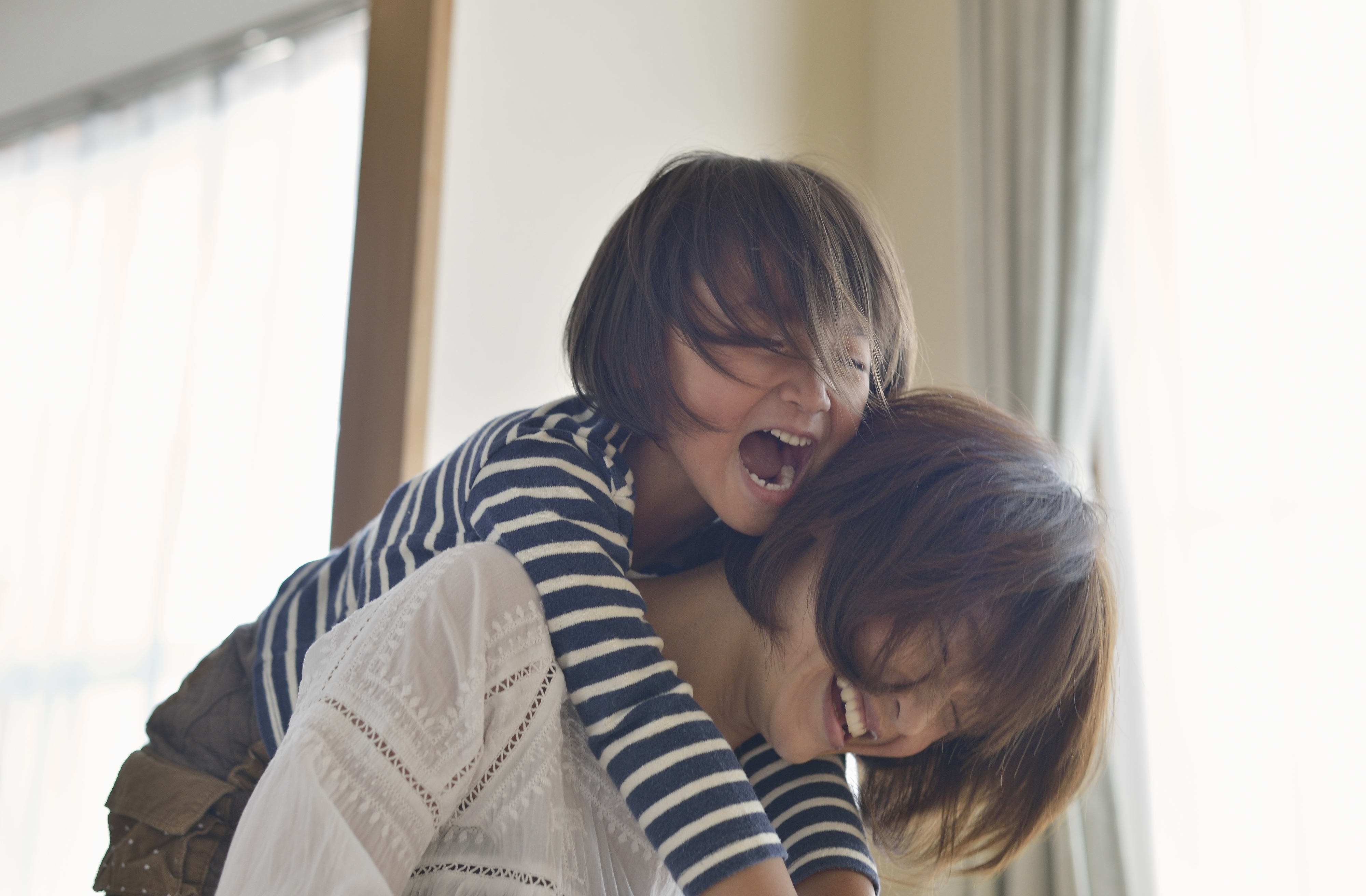 A smiling parent gives a piggyback ride to a laughing child wearing a striped shirt. Both have mid-length hair partially covering their faces
