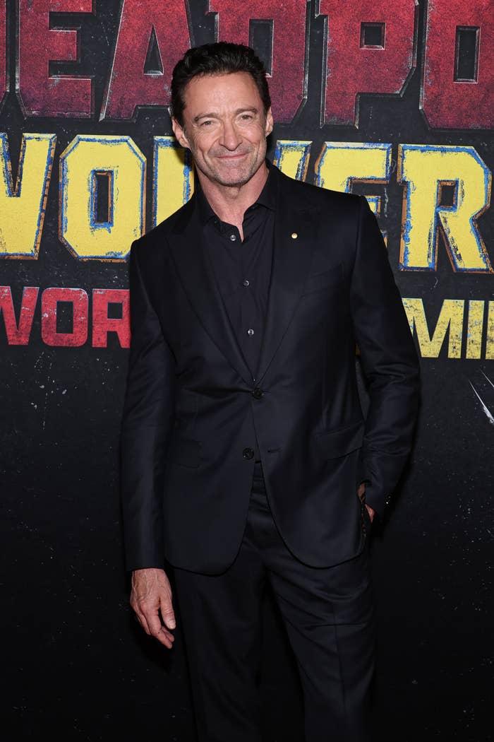 Hugh Jackman at a film event, wearing a black suit and smiling while standing in front of a &quot;Deadpool 3&quot; promotional background