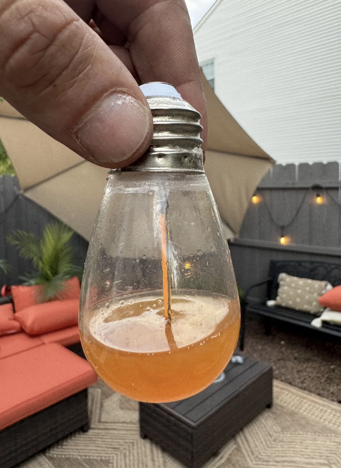 A hand holds a lightbulb filled with orange liquid over an outdoor patio with seating and an umbrella