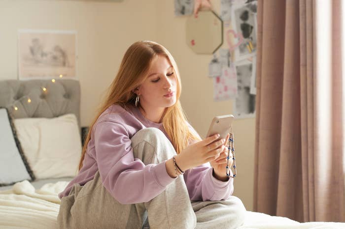 A person with long hair in a casual outfit, consisting of a sweater and sweatpants, is sitting on a bed looking at a smartphone. The room has a decorated wall