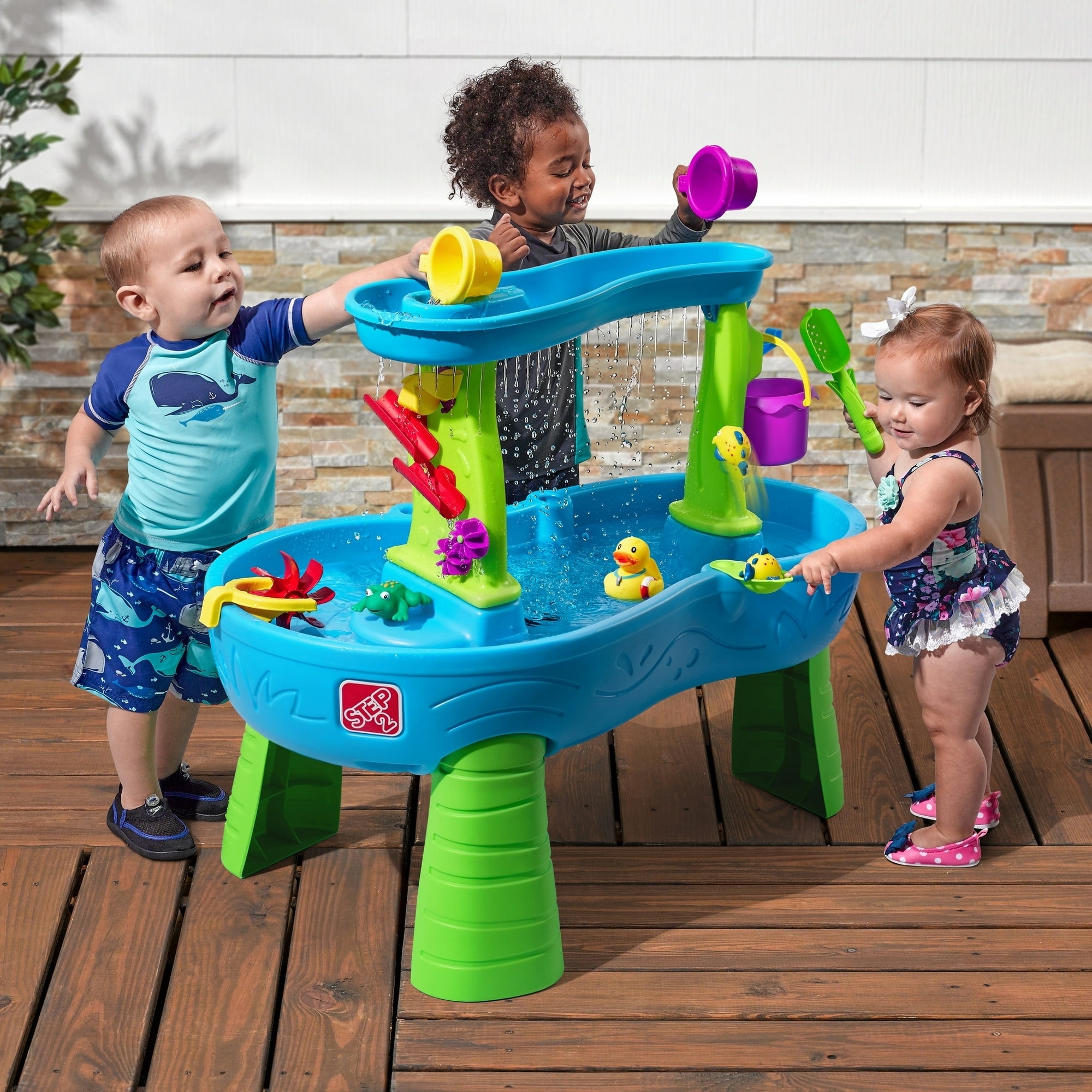 Three toddlers play with a colorful water activity table, pouring water and interacting with floating toys. The setting is outdoors on a wooden deck