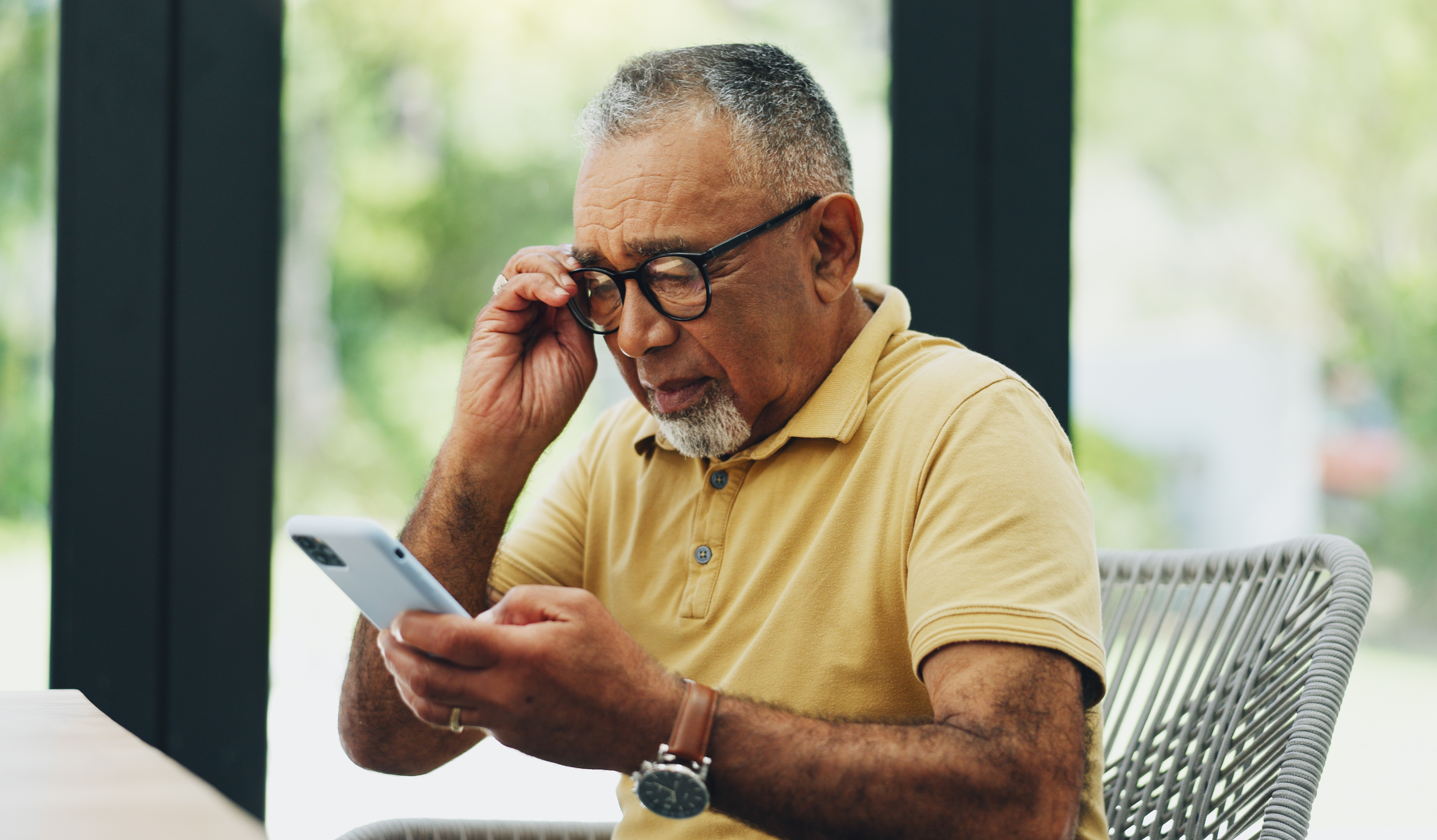 An older man with glasses wearing a short-sleeved collared shirt holds and looks intently at his smartphone while adjusting his glasses