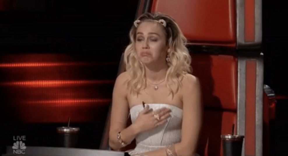 Miley Cyrus, in a strapless dress, sits in a red chair with a pensive expression and her hand on her chest, on the set of "The Voice."