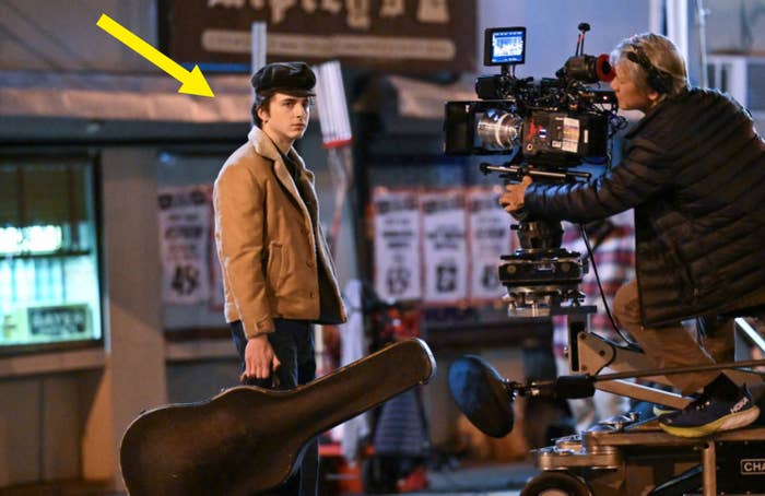 Timothée Chalamet holds a guitar case while standing next to a cameraman on a movie set