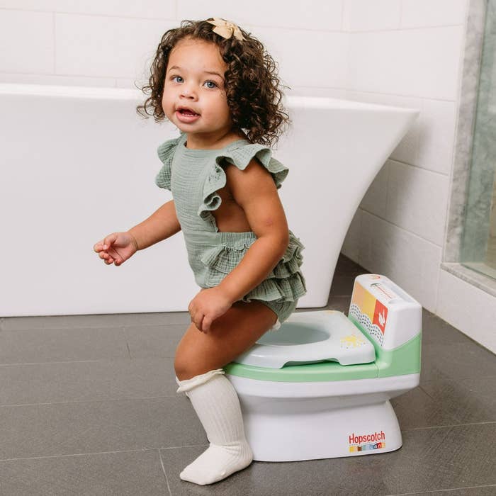 A young child, wearing a green ruffled outfit and knee-high socks, sitting on a children's potty labeled "Hopscotch." Backdrop includes a bathtub and tiled floor