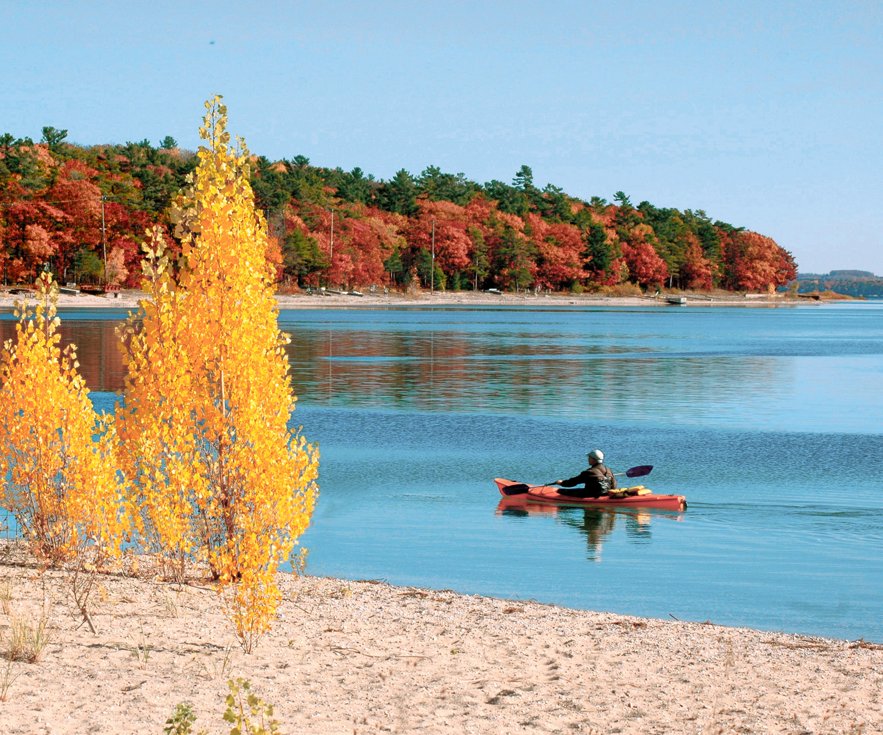 A person in a kayak paddles on a calm lake near a sandy shore with trees showing fall foliage in the background