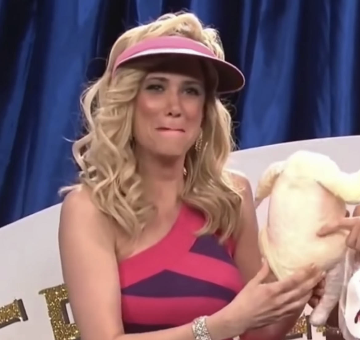 Kristen Wiig, in a pink and purple striped dress with a visor, holds a raw chicken while smiling at an event