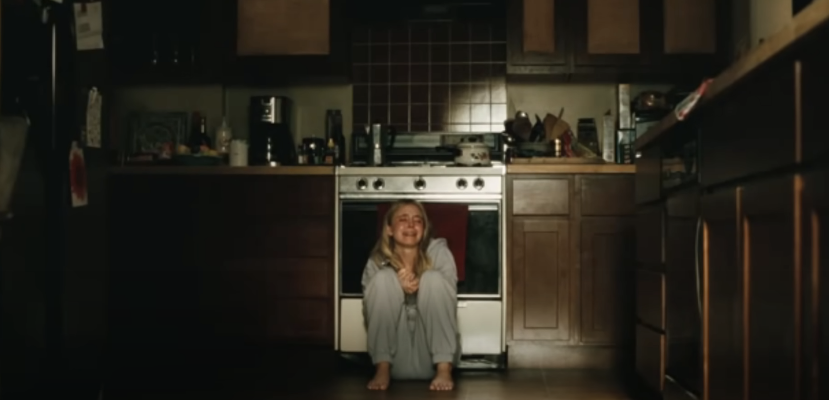 A person sits on a kitchen floor, smiling with knees hugged to their chest. The kitchen has wooden cabinets, a stove, and various items on the counter