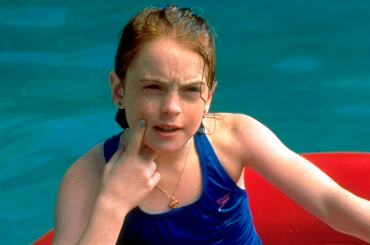 Lindsay Lohan in a swimming pool, wearing a blue swimsuit, touching her cheek with her finger, and looking thoughtful