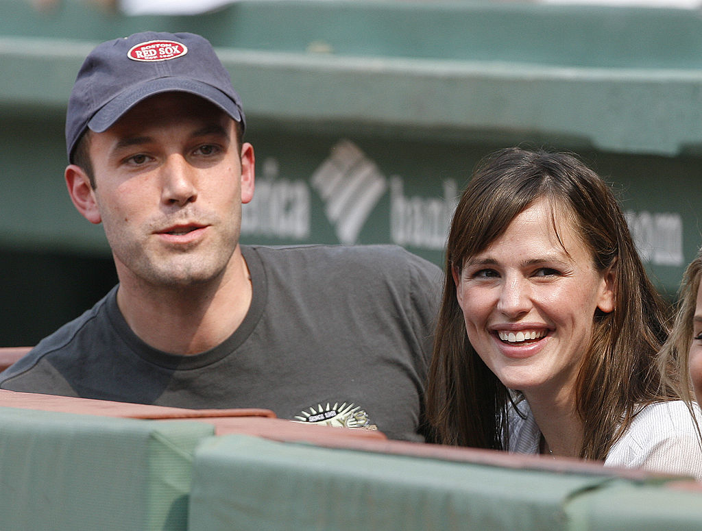 Ben Affleck and Jennifer Garner sitting in stadium seats, smiling and looking in the same direction. Ben wears a baseball cap and T-shirt