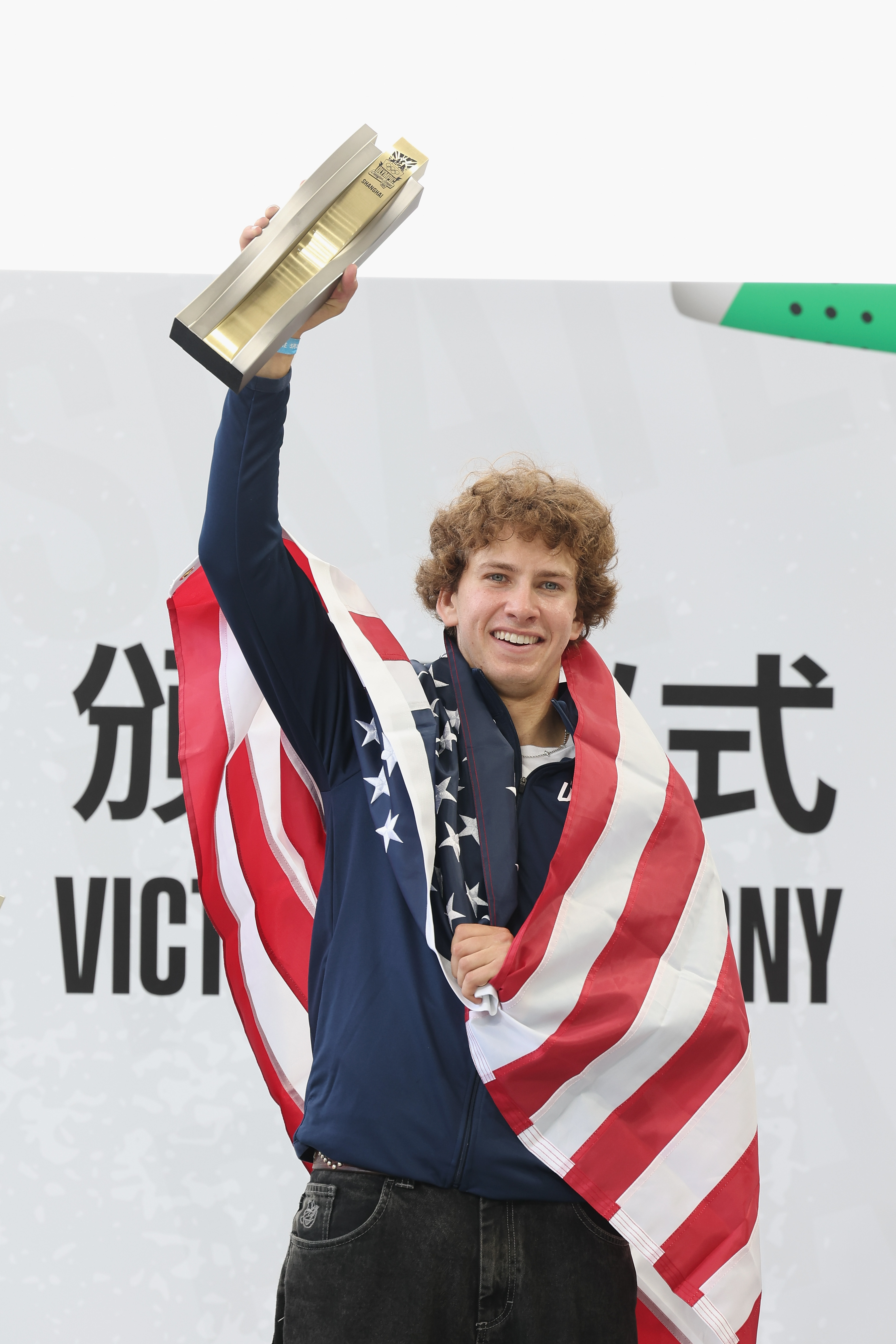 Athlete Nathan Chen draped in an American flag, holding a trophy above his head, smiles during a victory ceremony